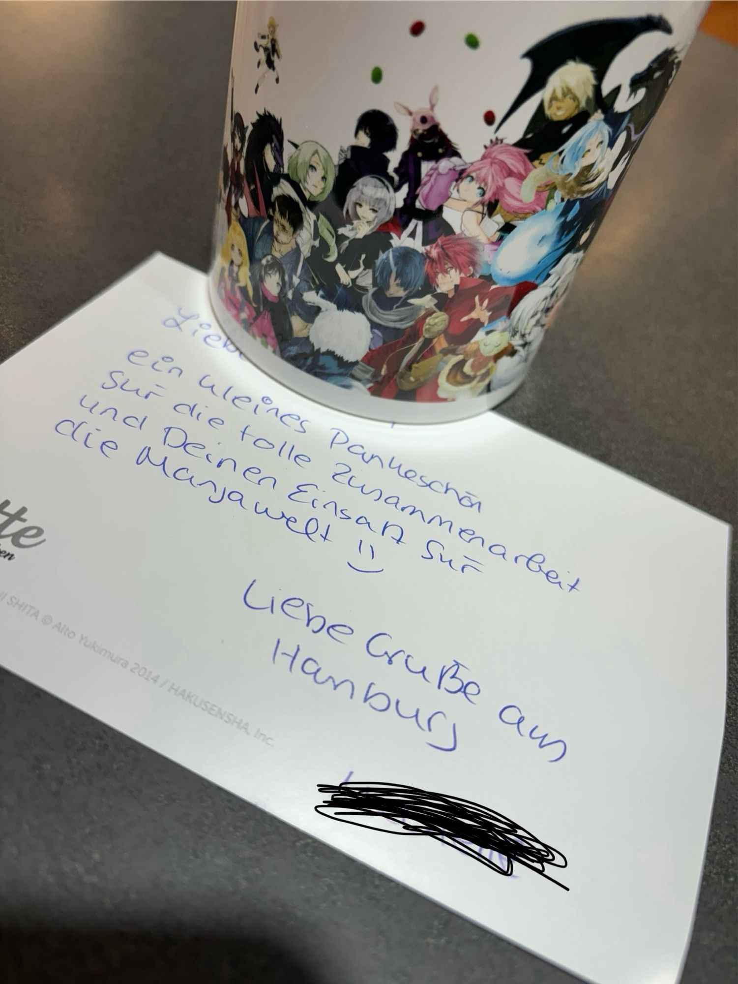 Eine Tasse mit Mangafiguren verziert und eine Postkarte auf einem Tisch, Text auf der Karte: „Lieber …, ein kleines Dankeschön für die tolle Zusammenarbeit und Deinen Einsatz für die Mangawelt 😃
Liebe Grüße aus Hamburg…“