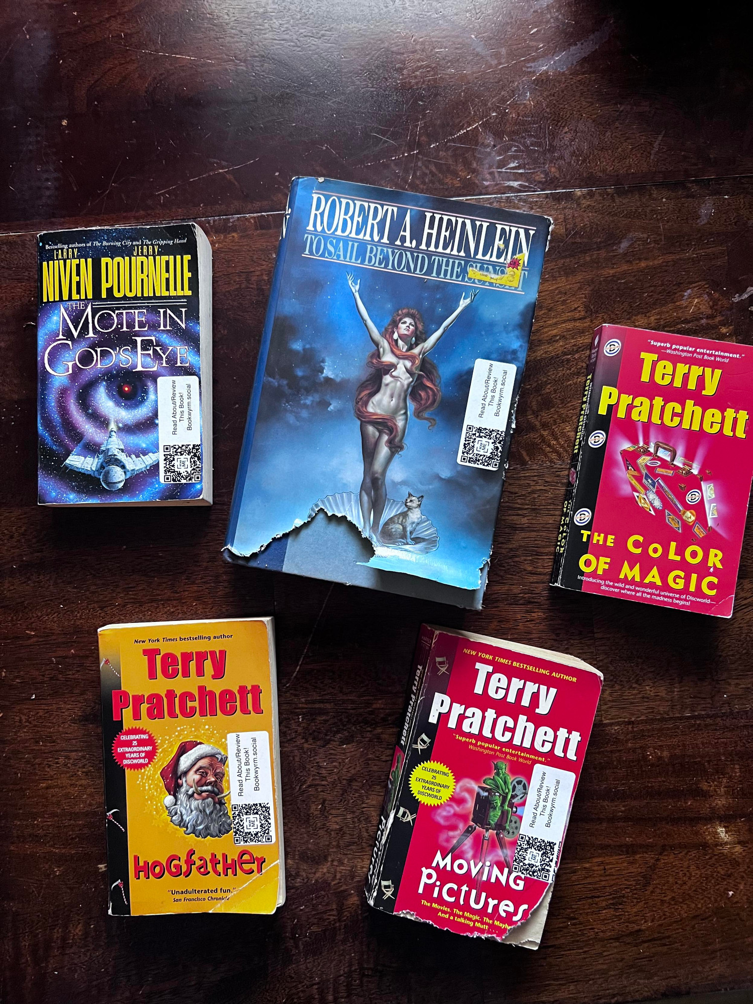 Picture of five books sitting on a wooden table, from left right, top to bottom: “the Mote in Gods Eye“ by Larry Niven and Jerry Pournelle, “To Sail Beyond the Sunset“, by Robert a Heinline, “The Color of Magic“, Hogfather“ “Moving Pictures“ all by Terry Pratchett