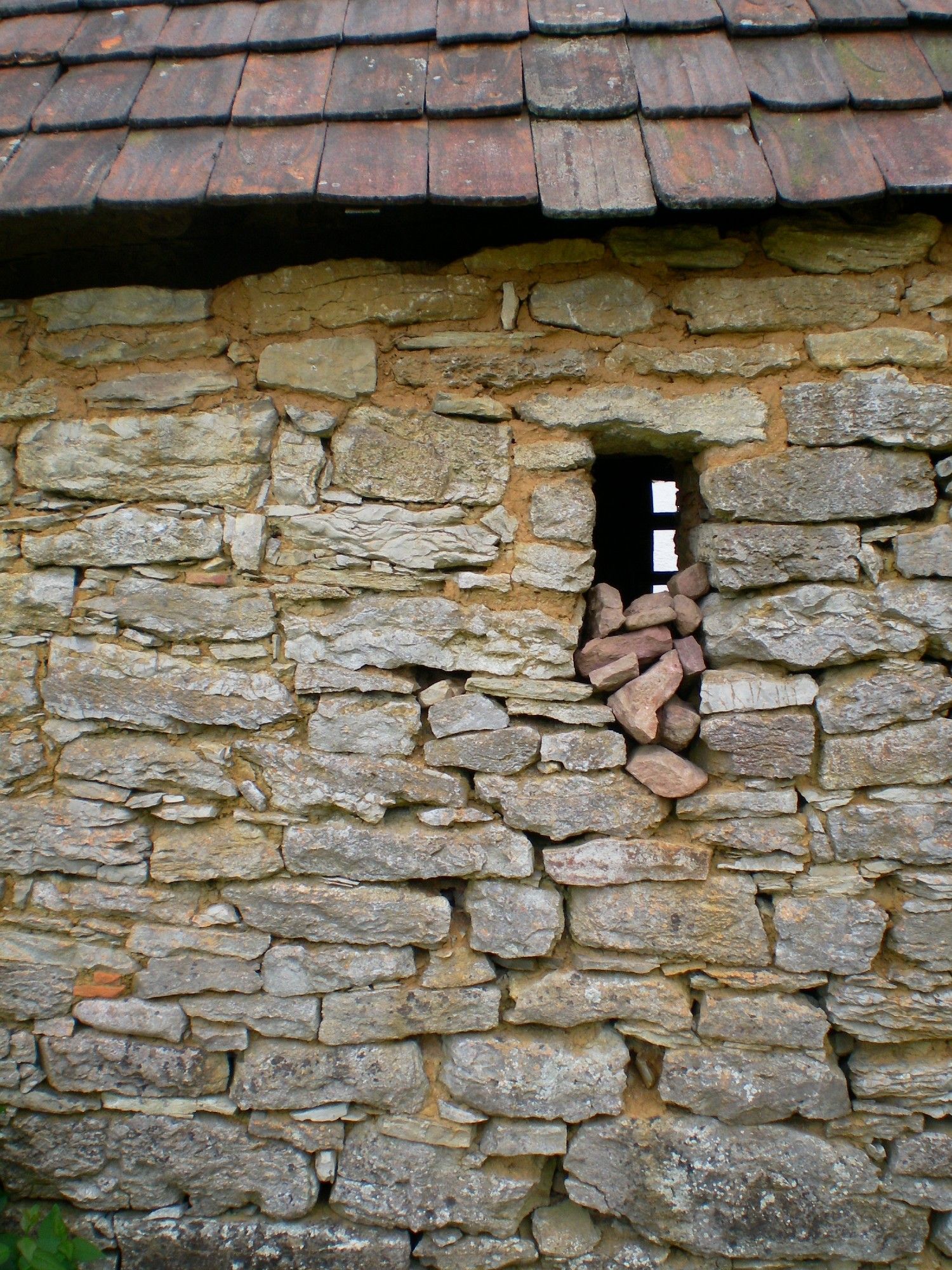 Das Bild zeigt eine aus verschieden großen Steinen sehr lose gemauerte Außenwand, in der ein schießscharten-ähnliches Fenster zu sehen ist welches nach unten hin beschädigt zu sein scheint. Man hat wohl kleine Steine vom Weg hineingelegt um den Schaden zu füllen. Terracottafarbene Dachschindeln am oberen Bildrand weisen darauf hin, dass es ein altes, Landwirtschaftsgebäude sein könnte.