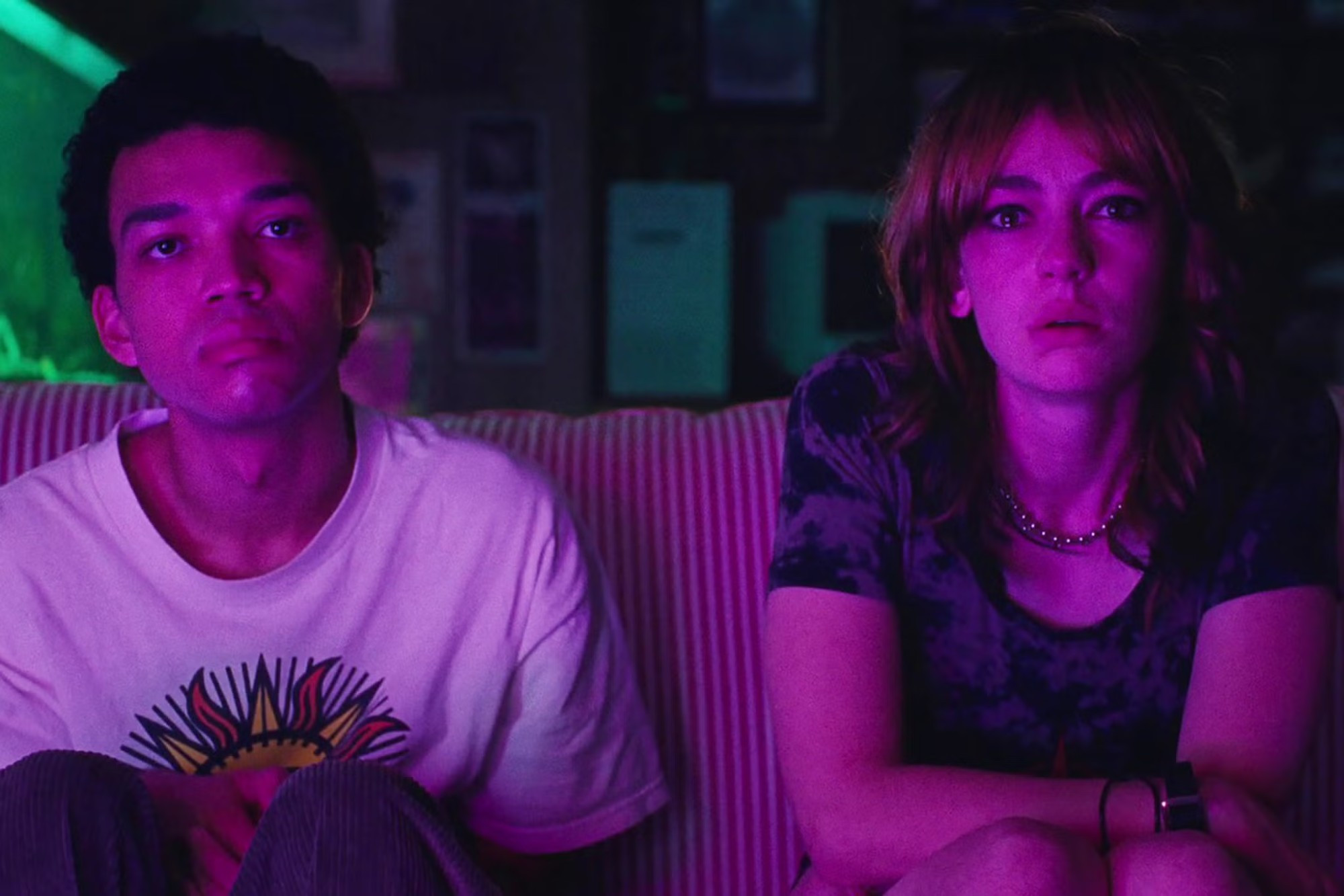 Justice Smith and Brigette Lundy-Paine as Owen and Maddy in “I Saw the TV Glow”; the two are sat on a sofa, bathed in pink light from the TV they are watching (out of shot)