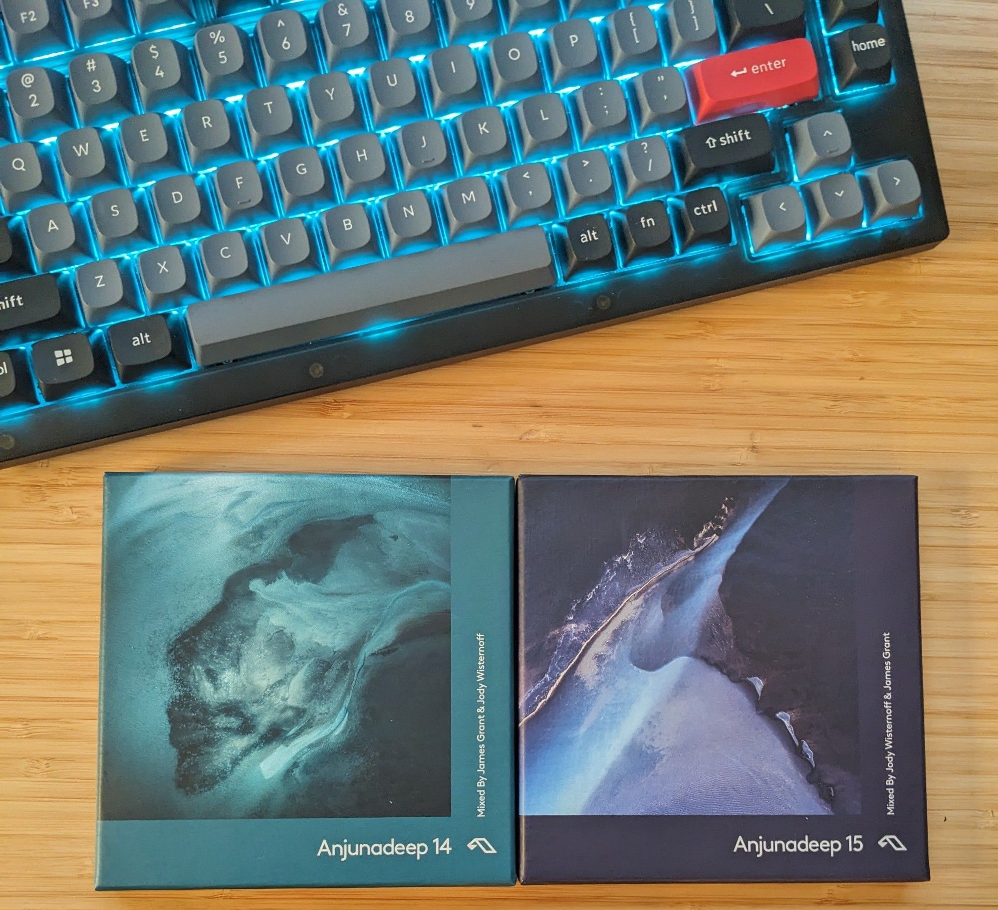 Physical copies of Anjunadeep 14 and 15, organized side by side on a bamboo desk. A keyboard with aqua lighting is positioned above.