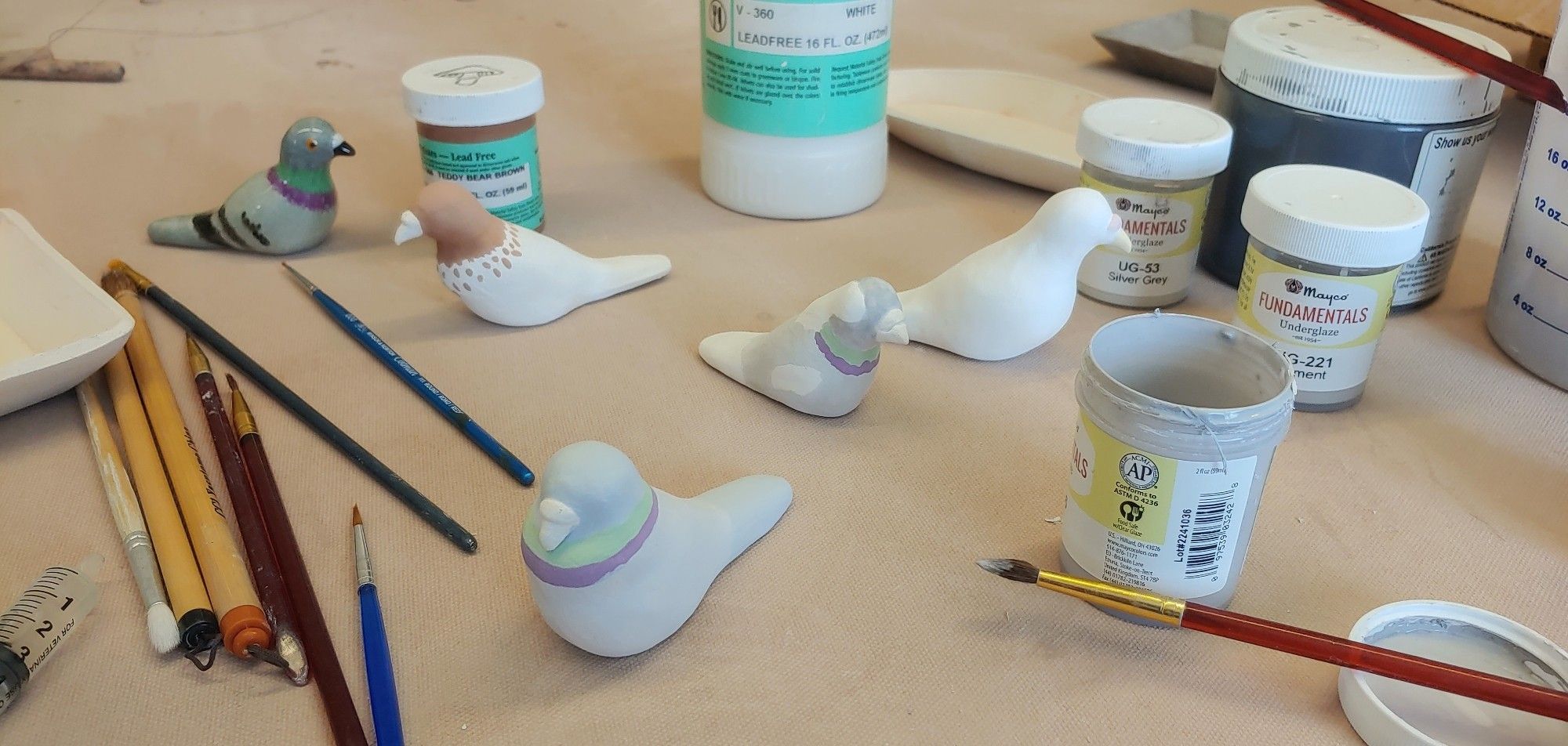 Four ceramic pigeons getting underglazed with paintbushes and underglazes surrounding them. In the back is a finished pigeon.