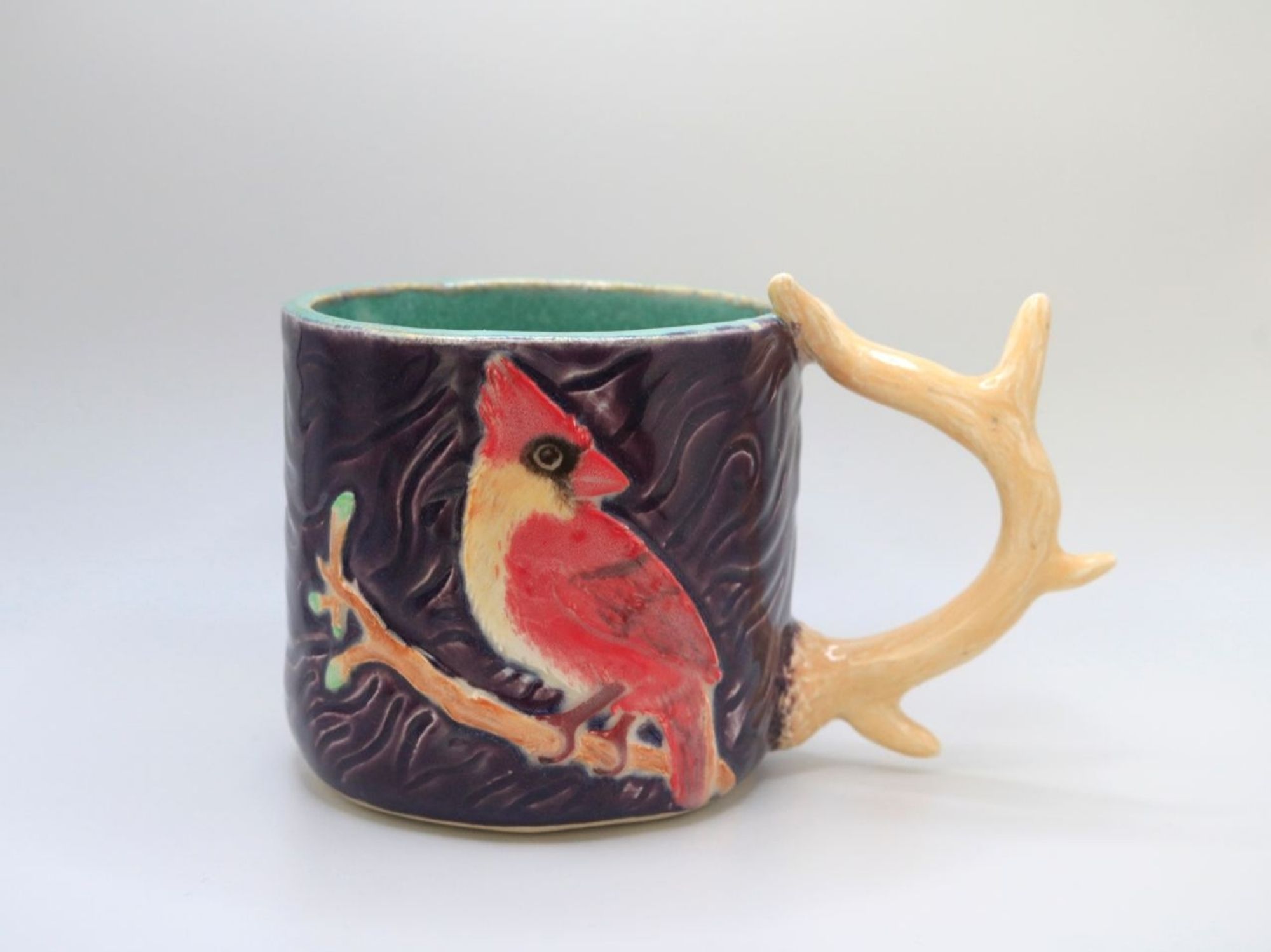 A ceramic mug with a gynandromorphic cardinal perched on a branch carved into the side. The mug has an antler shaped handle.