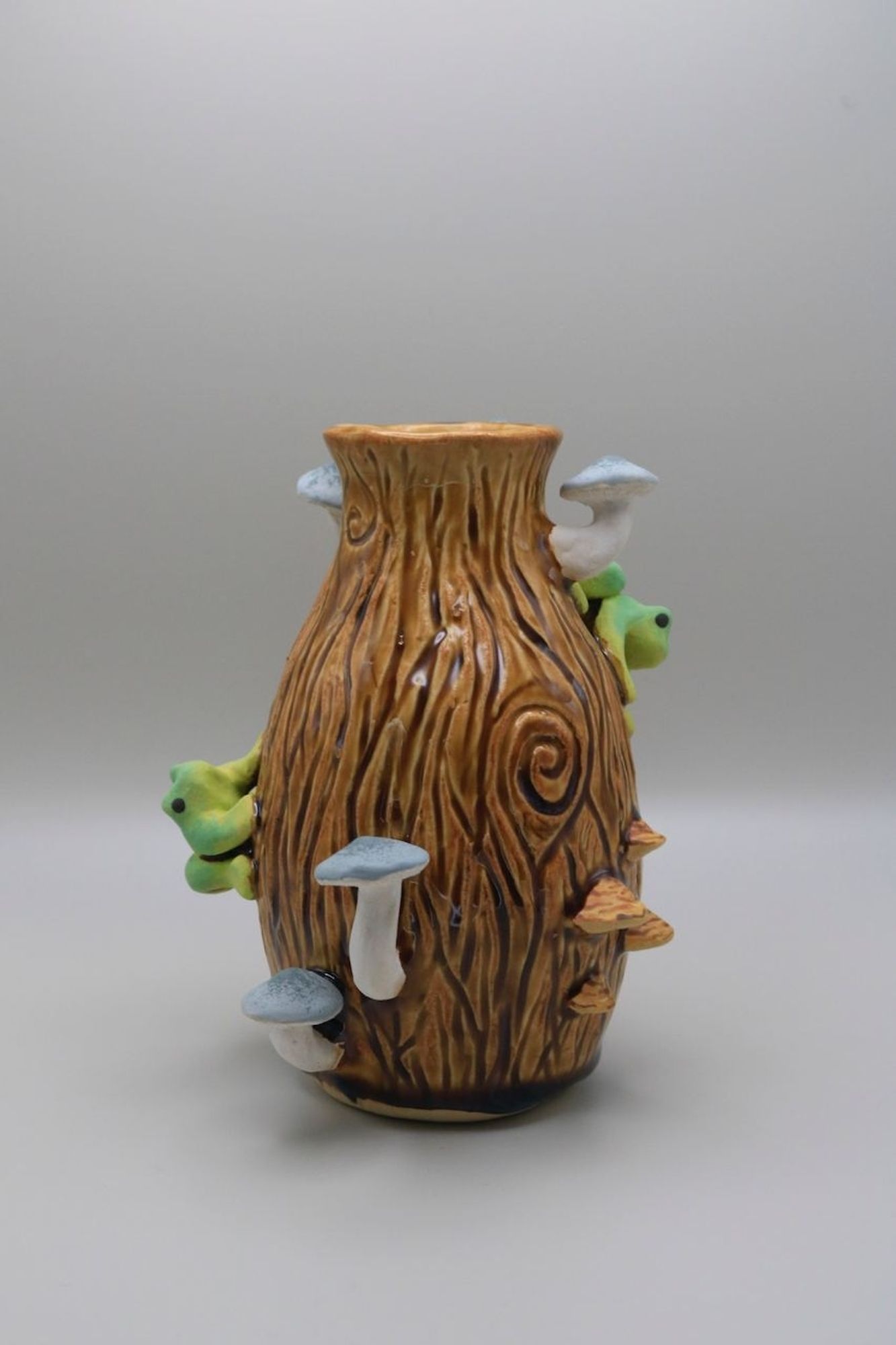 A ceramic vase textured like wood with mushrooms and frogs sculpted on it throughout.