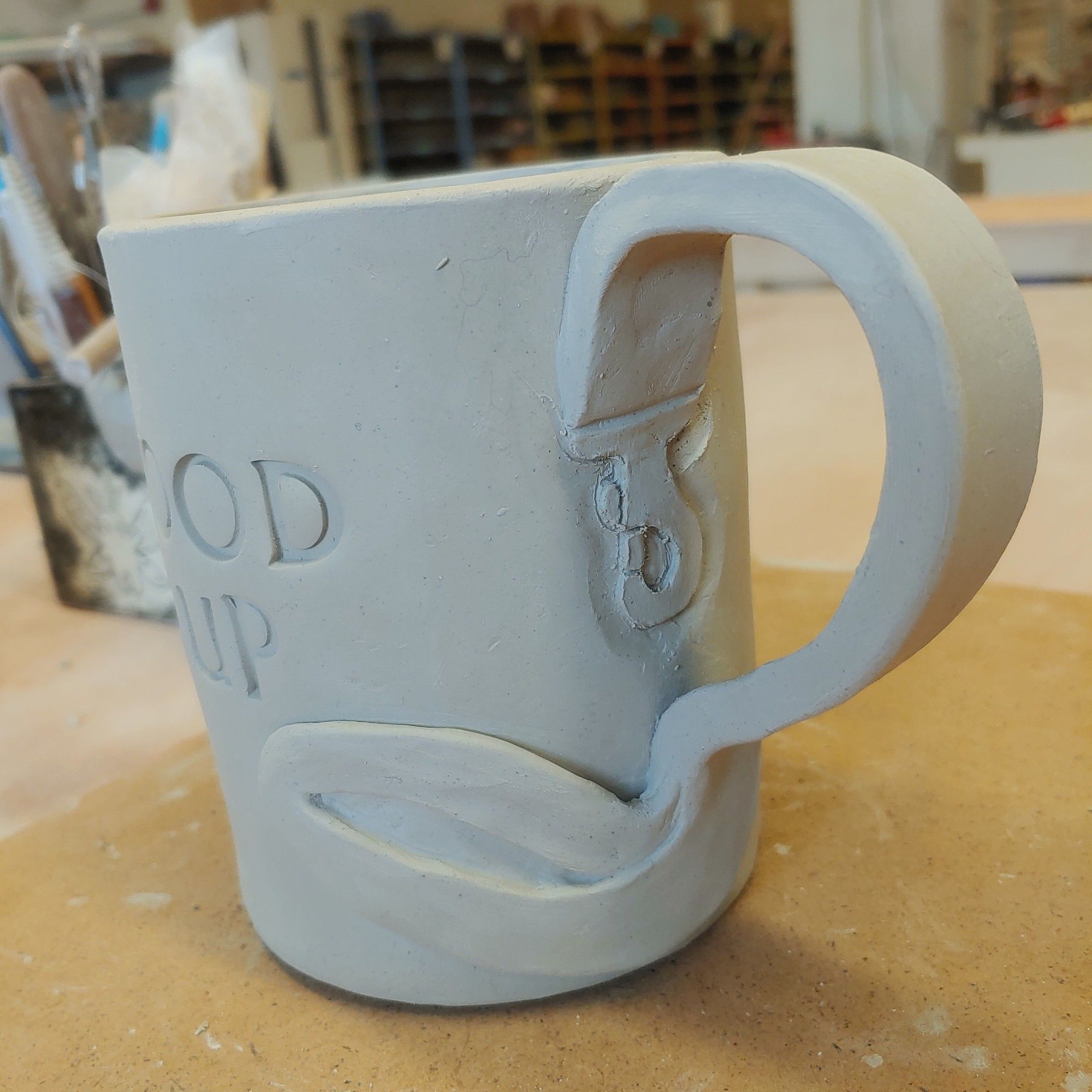 A ceramic mug in the greenware stage the handle is in the shape of a leash and on the side words are pressed in the phase is cut off the the letters seen are "od" and "up"