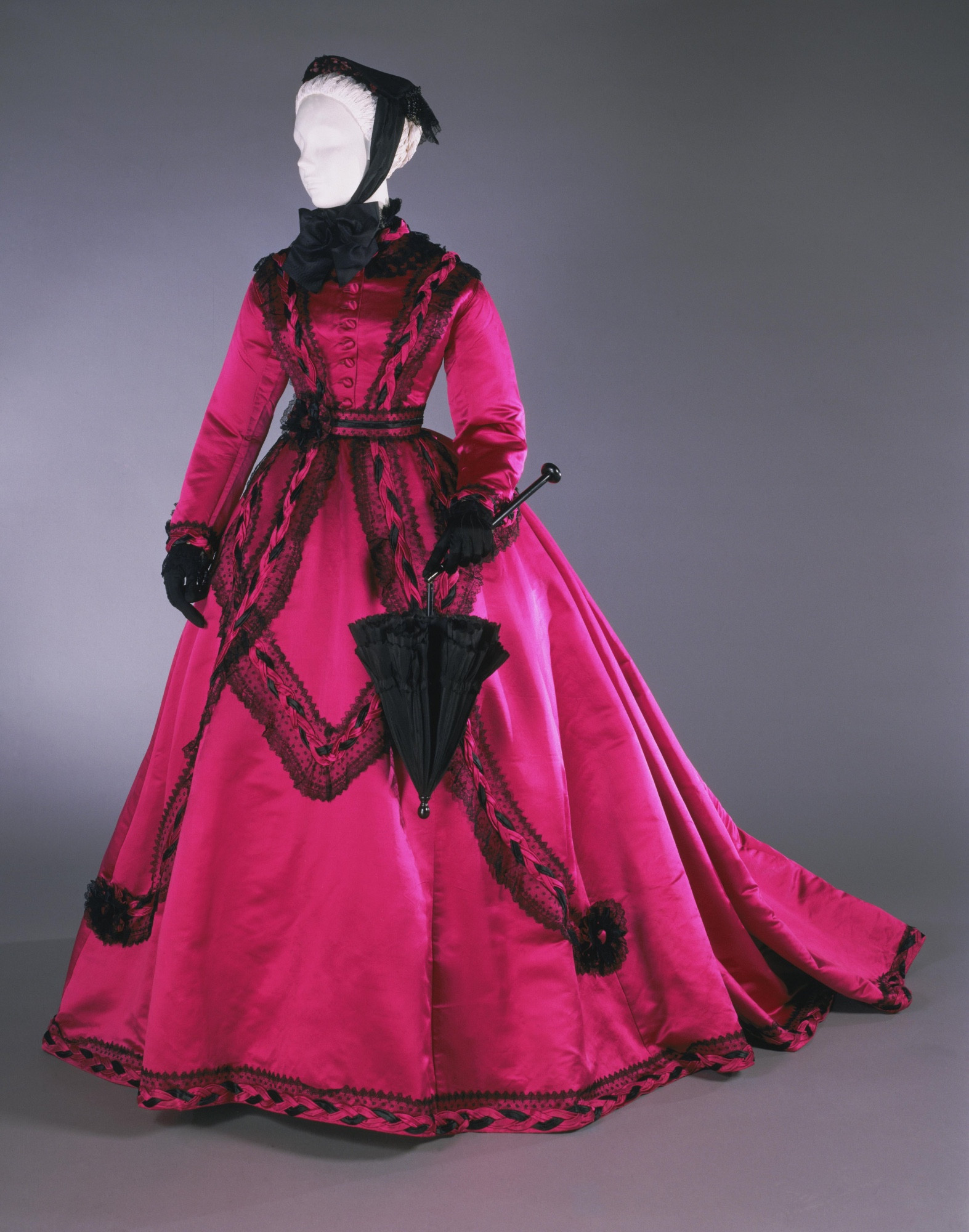 A brilliant magenta dress of silk with black lace trim edges and details. Conical sweeping skirt of era. The bodice has long sleeves. 