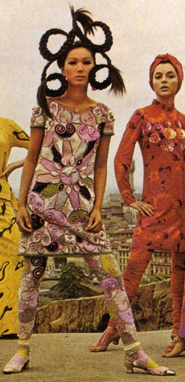 Two models wearing sixties fashion - dresses and leggings with bold graphic prints, one in pink-green-white colors another one in reddish hues. One of the women has an outlandish hairstyle consisting of braids framed in looping circles 