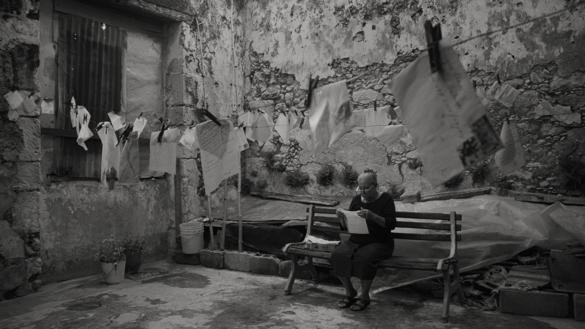 An old lady sits on a bench with a letter, lots of other letters are pegged to a washing line in front of her. Still from Oceans Are The Real Continents