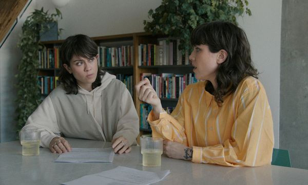 A still from the film Fanatical: The Catfishing Of Tegan And Sara. Two white women, identical twins with dark shoulder-length hair and concerned expressions, sit at a table, talking to each other, with document and drinks in front of them. Behind them are crowded bookshelves with thriving plants on top of them.