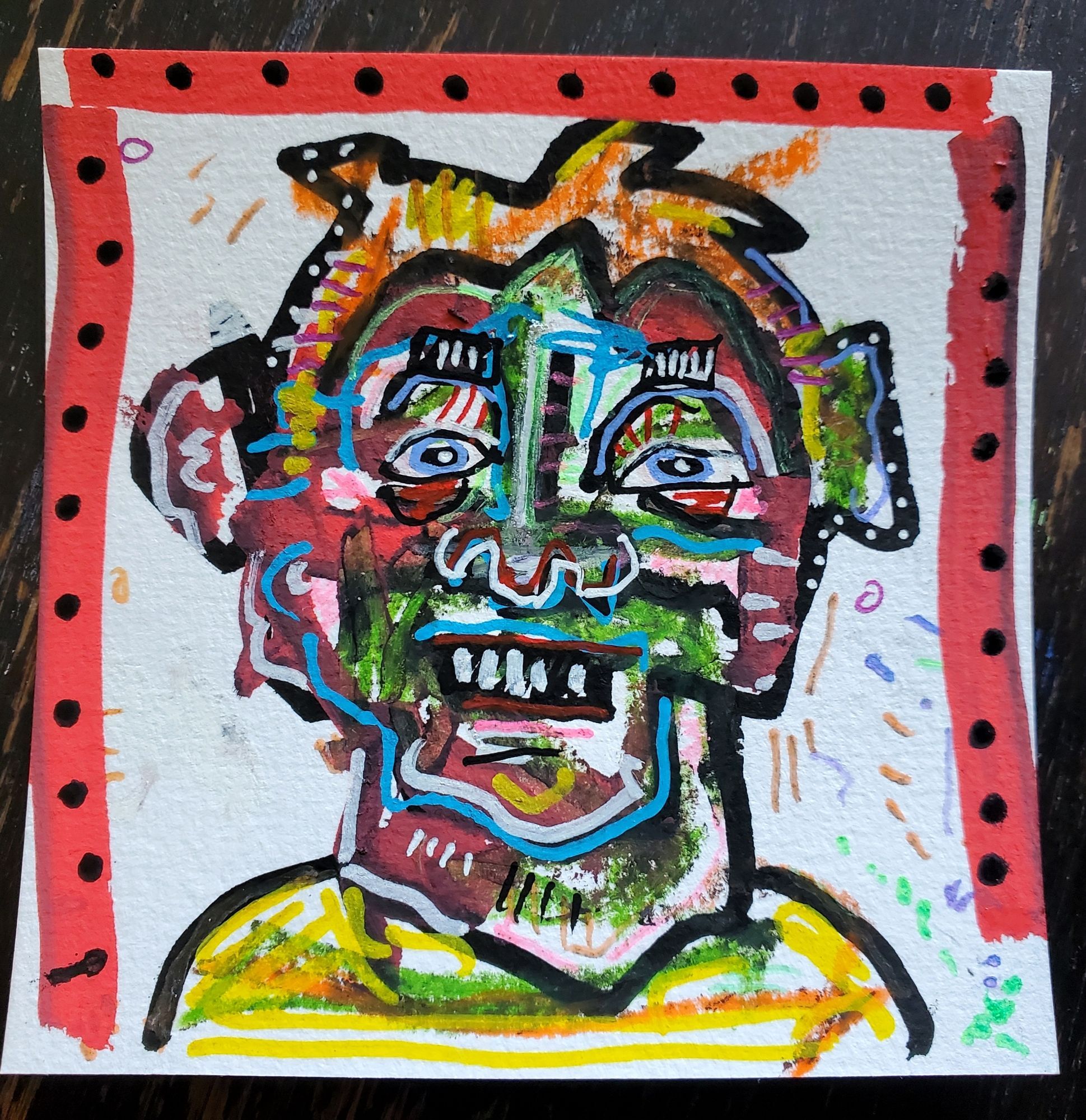 A small card with a face drawn on it. Paint pens and other media. The person is grinning like an idiot and is missing a tooth. Sloppy.