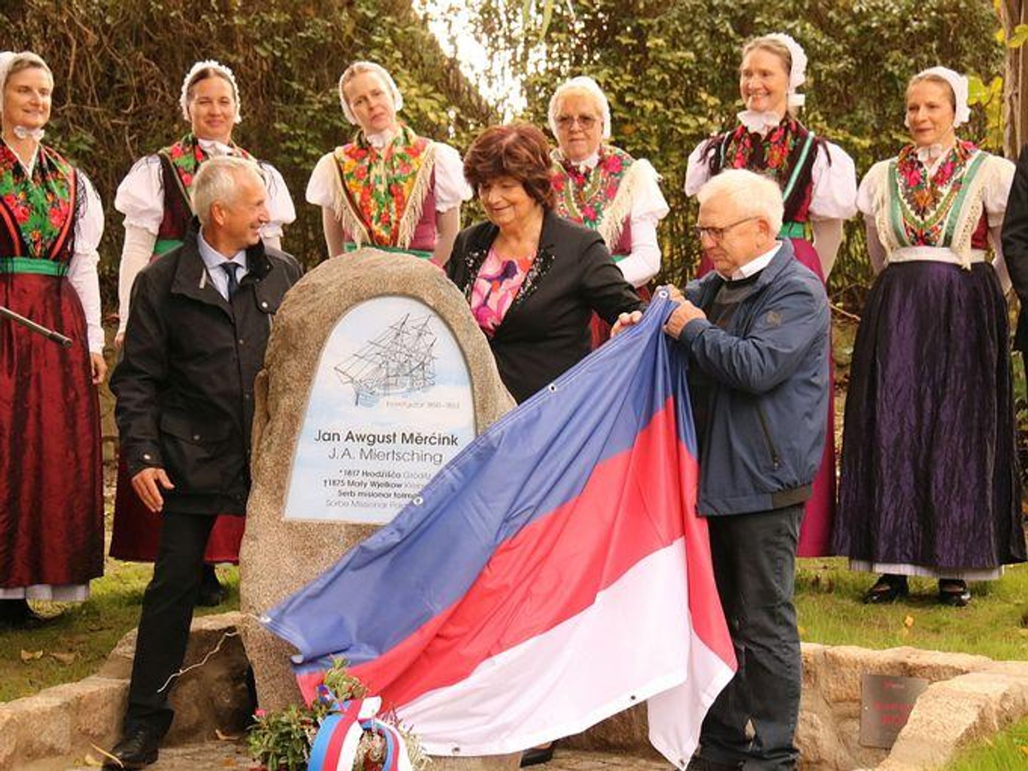 Auf dem Foto stehen Frauen in sorbischer Tracht, davor ein Gedenkstein, von dem gerade eine sorbische Flagge entfernt wird, die ihn zuvor noch verhüllt hatte. 