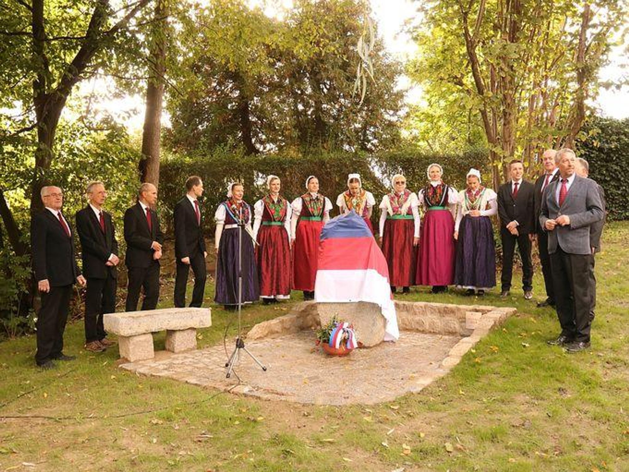 
Auf dem Foto singt ein gemischer Chor mit Frauen in sorbischer Tracht unter großen Bäumen vor einem mit einer sorbischen Flagge verhüllten Monument, neben dem eine steinerne Bank steht. 