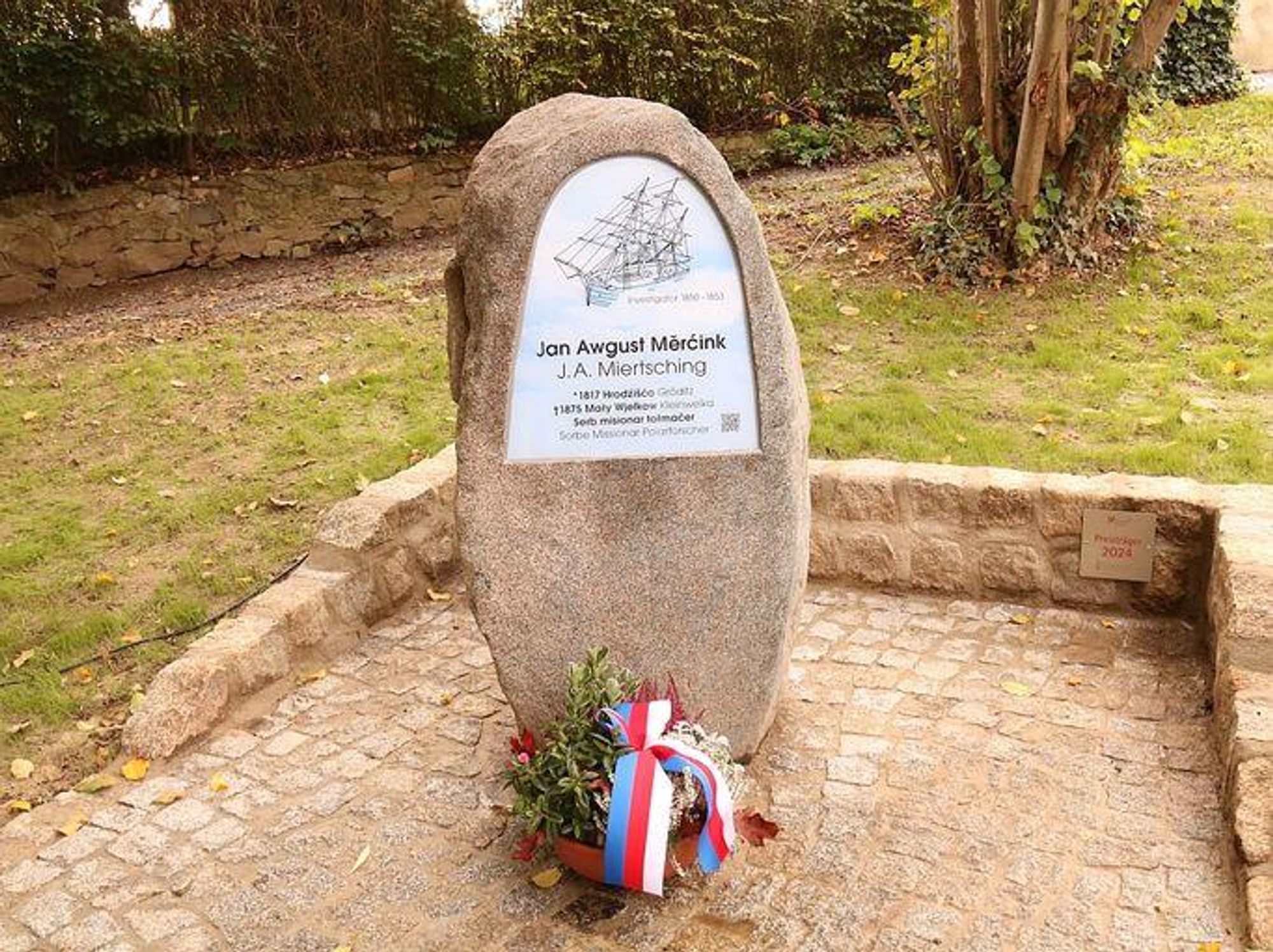 Das Bild zeigt ein Denkmal - einen Findling, an den eine Tafel montiert ist, die oben die silhuette eines Schiffs zeigtm darunter Namen und Daten der geehrten Person Johann August Miertsching in sorbischer und deutscher Sprache. 