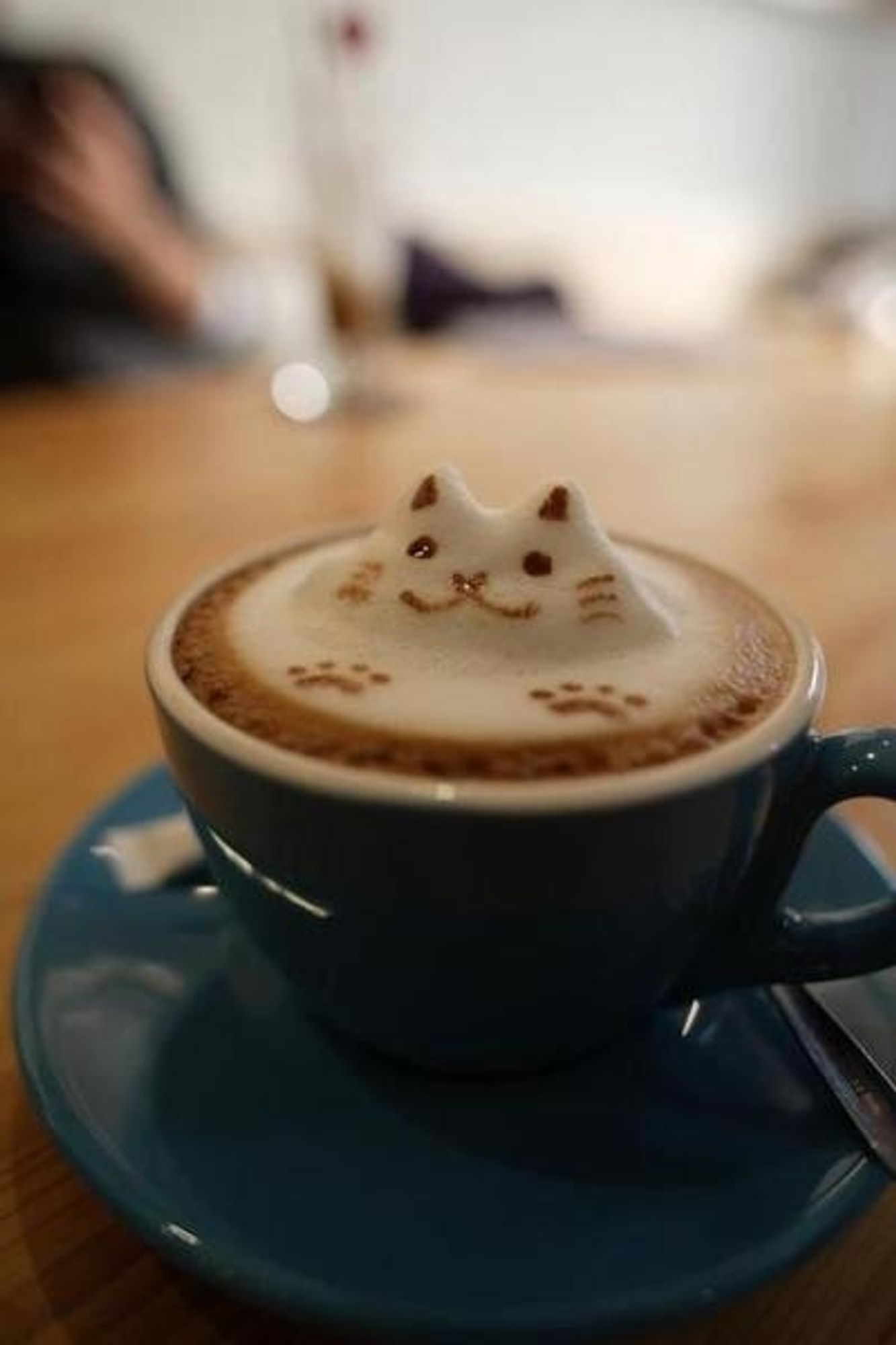 A blue ceramic coffee cup on a matching saucer. The coffee has a foam layer on top that is decorated to look like a cute little kitty