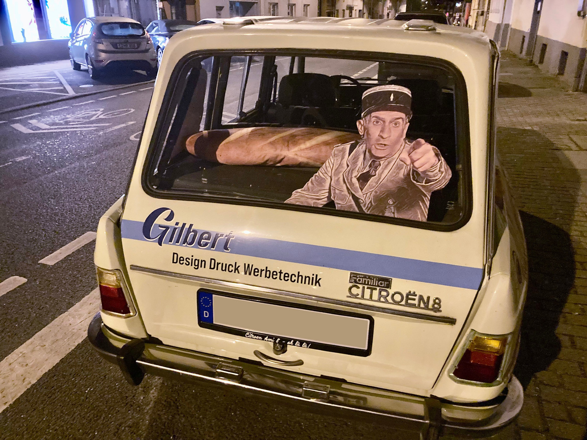 Photo de l'arrière d'une Citroën Ami 8 garée à Essen en Allemagne. Elle est décorée d'une photo géante du gendarme de Saint-Tropez (Louis de Funès) sur la vitre arrière. Sur la plage arrière, on aperçoit un coussin en forme de baguette de pain géante.