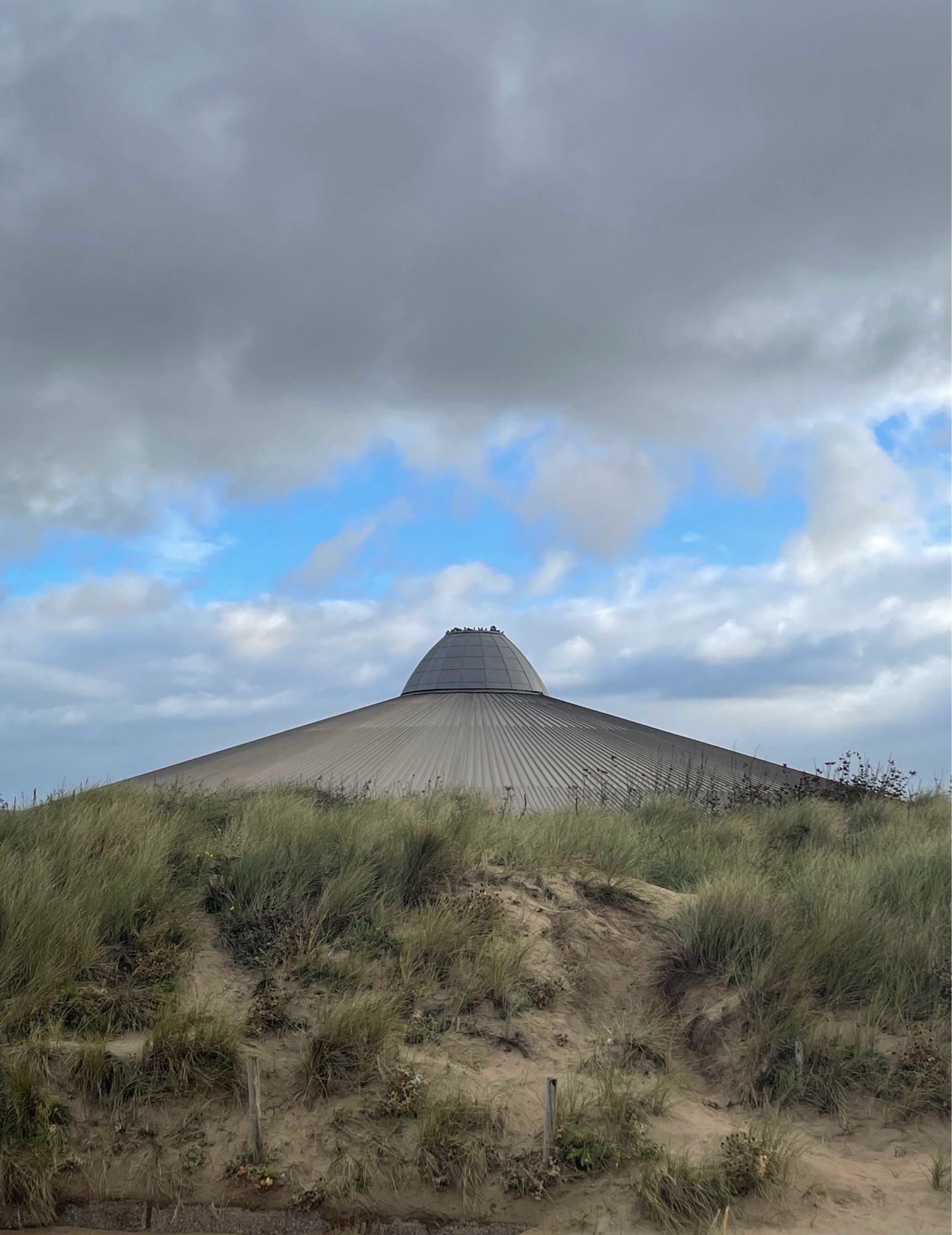 Four photos of a building shaped like a 1950s B movie UFO, facing onto sand dunes. There are pigeons on top of the building.