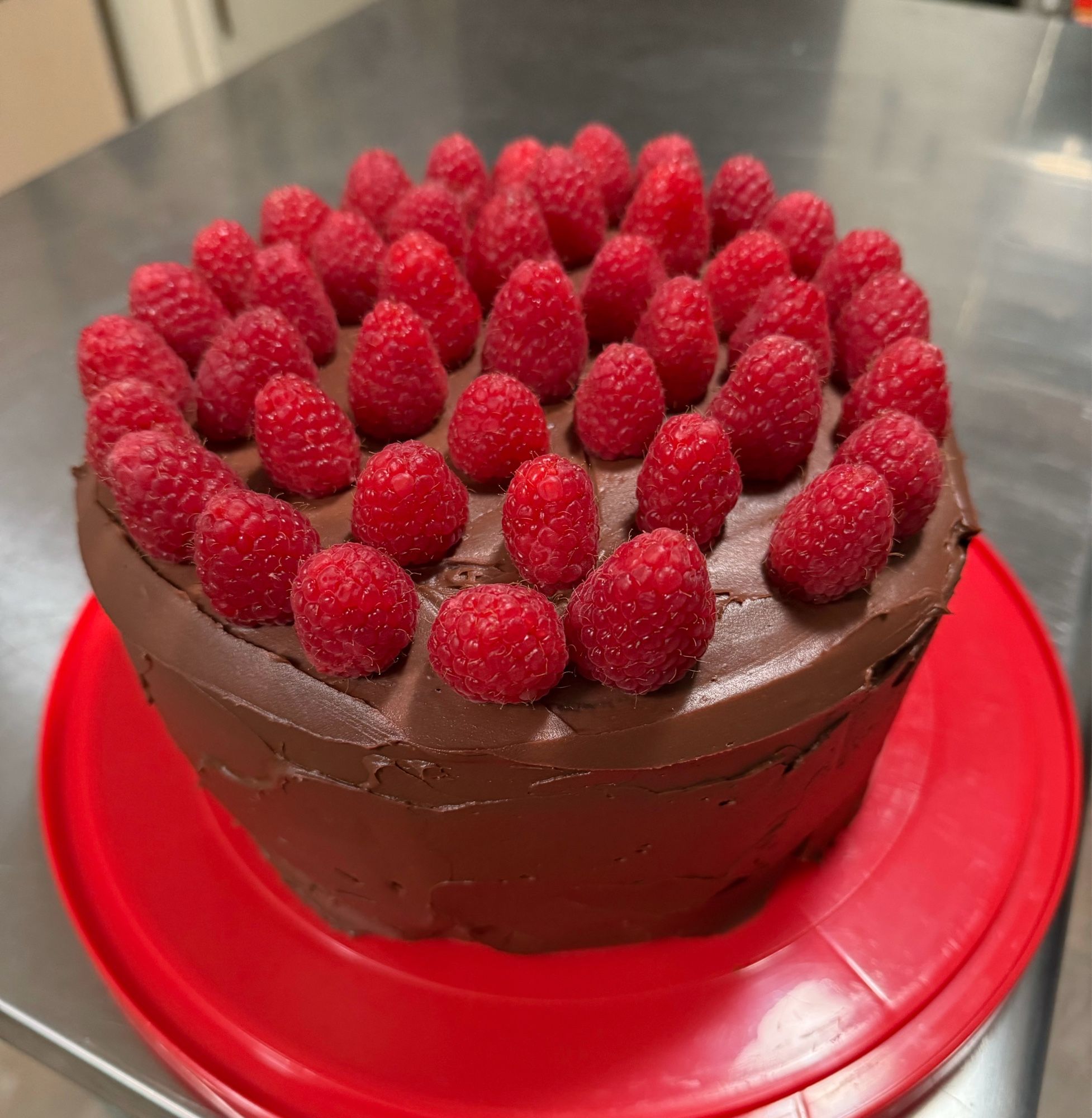 Two tier chocolate cake with chocolate ganache frosting. It is topped with raspberries and there is a raspberry missing from the outer edge.