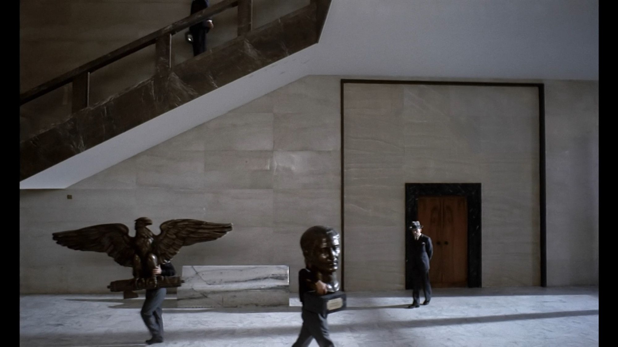 Screen from "The Conformist" with the protagonist waiting on the ground floor of a white marble building as workers go past carrying busts of fascist symbols.