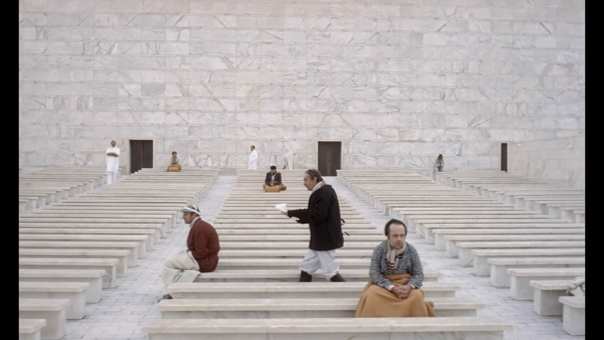 Screen from "The Conformist" with people in an asylum made of white marble, the room filled with rows of low benches that the patients sit on.