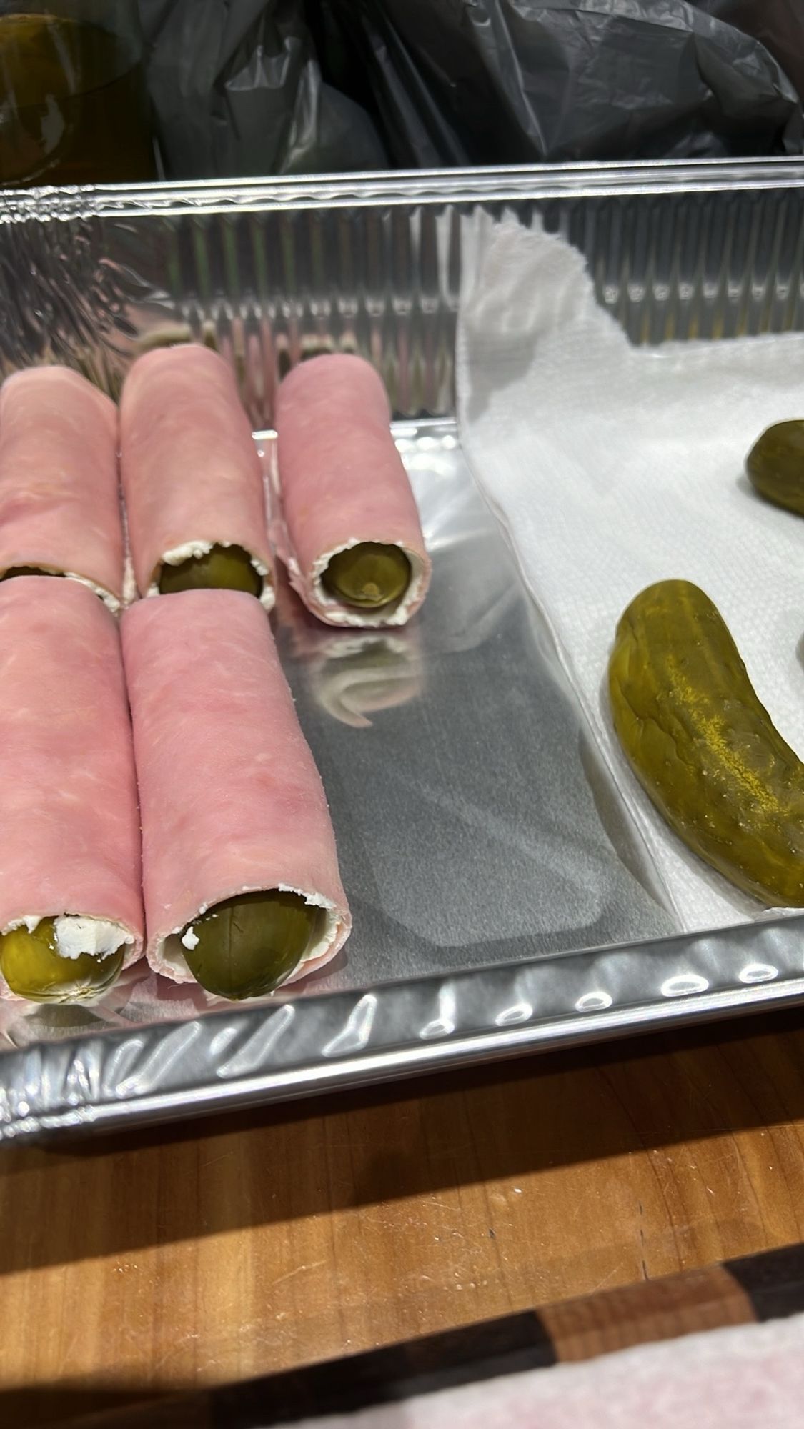 Five pregnant pickles, which is deli ham with cream cheese on it and wrapped around a whole dill pickle, in a food prep pan with other pickles in the pan waiting to be finished