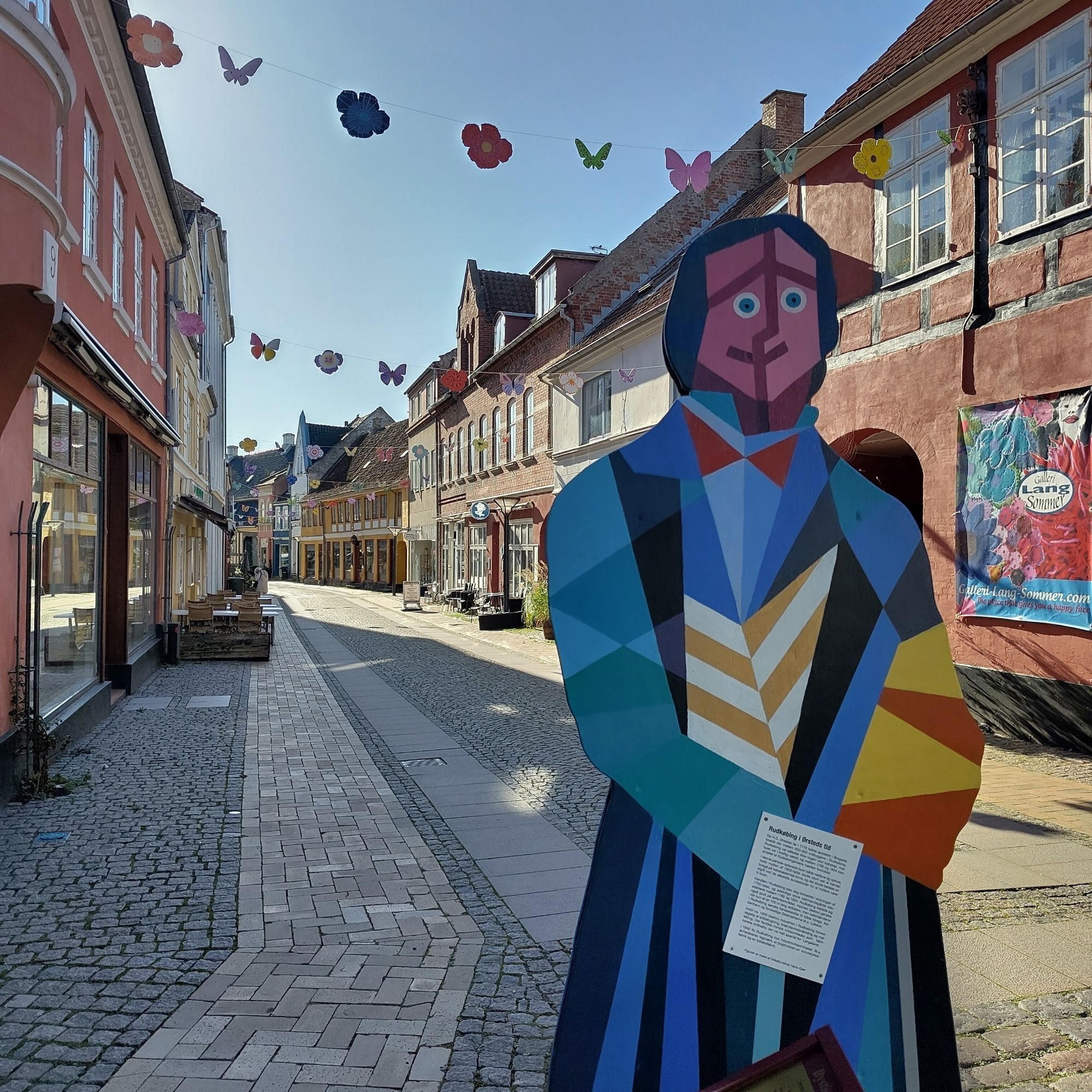 Eine leere Fußgängerzone, im Vordergrund eine bunt bemalte Skulptur. Über der Straße hängen bunte Blumen und Schmetterlinge.