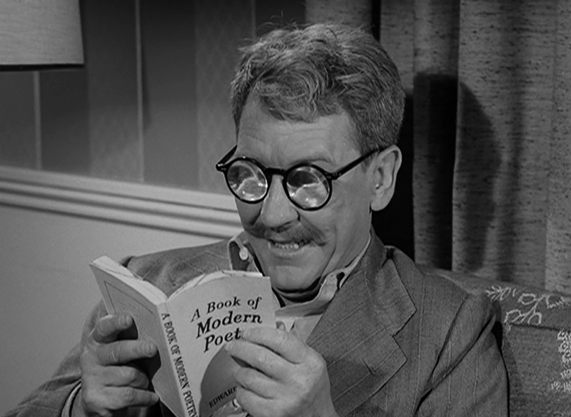 A man with very think glasses reads from a book of modern poetry. It is Henry Bemis from The Twilight Zone.