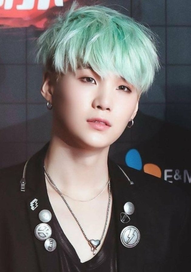 Yoongi is standing and looking forward while wearing a black blazer with some pins and buttons on it and a thin black shiny blouse underneath it. He has messy light green hair and a very light smoky eye makeup. He's also wearing earrings and a necklace with a melting heart pendant. This photo is probably from 2015, Run era.