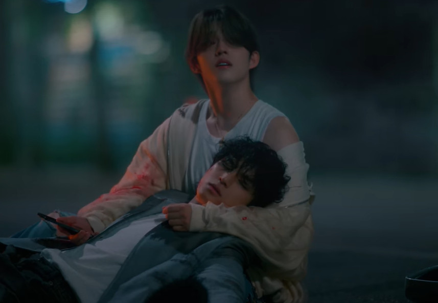 screenshot from the 'eyes on you' mv by kpop boy group seventeen, seungcheol (scoups) cradling mingyu in his arms, they're both on the road and there is blood on their clothes