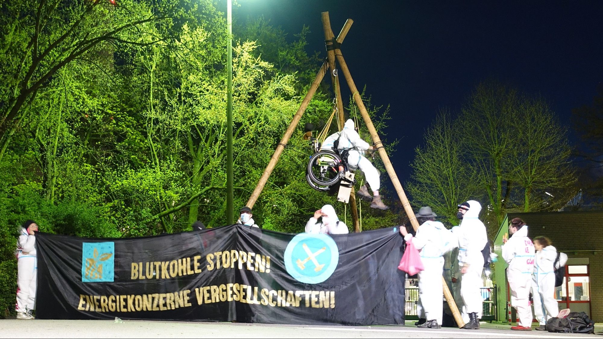 2 Menschen hängen in einem Dreibein , eine der 2 Personen im Rolltuhl. es ist Nacht. die Demonstrierenden tragen weiße Anzüge. auf dem Banner vor dem Tripod steht Blutkohle stoppen Energiekonzerne vergesellschaften und das Logo von Ende Gelände