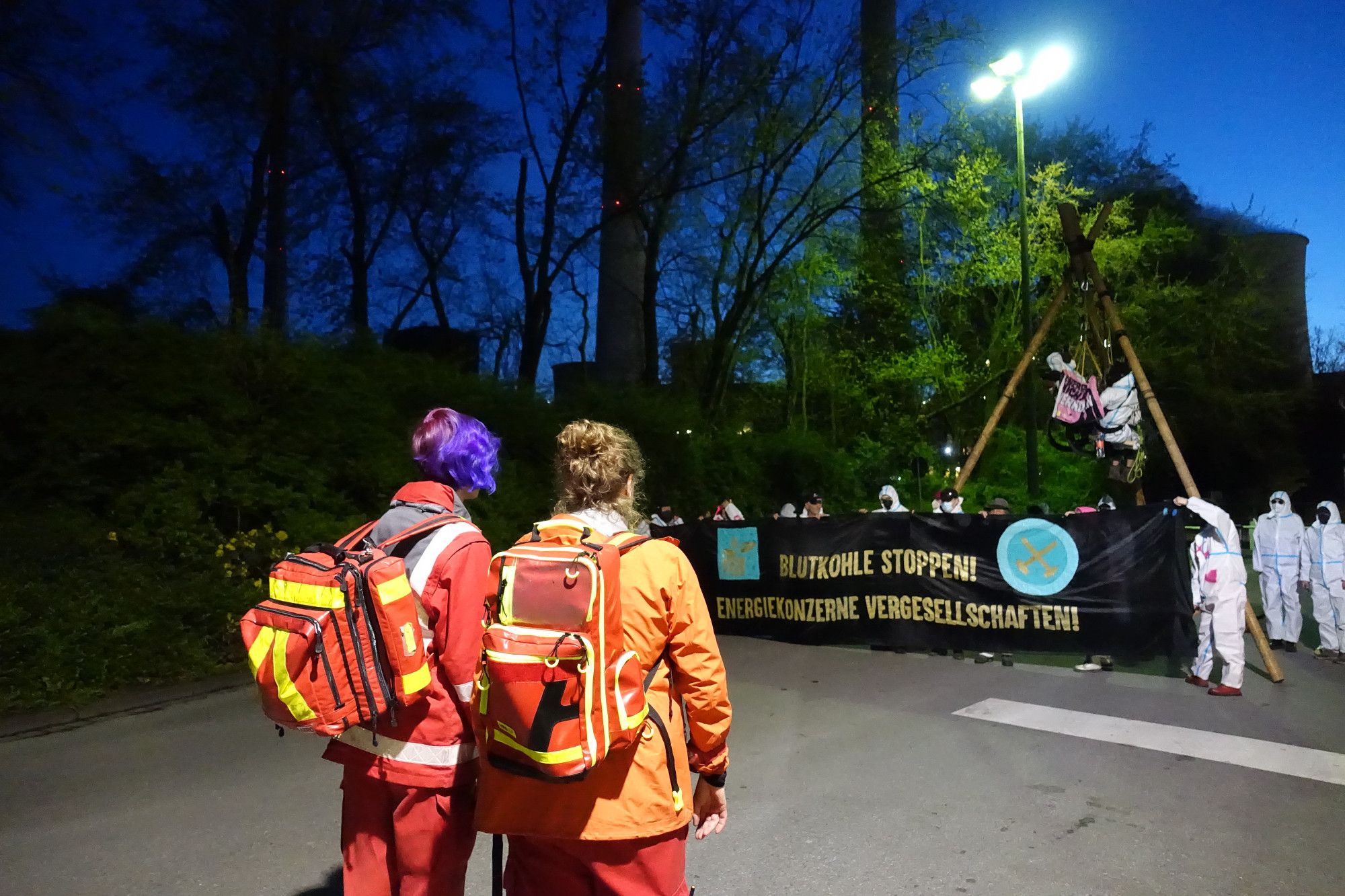 2 Demosanitäter*innen mit Oranger weste stehen vor der Tripod-Aktion. Im Hintergrund die Aktion mit Tripod, 2 Menschen darin, Menschen mit weißen Maleranzügen und banner Blutkohle stoppen energiekonzerne vergesellschaften