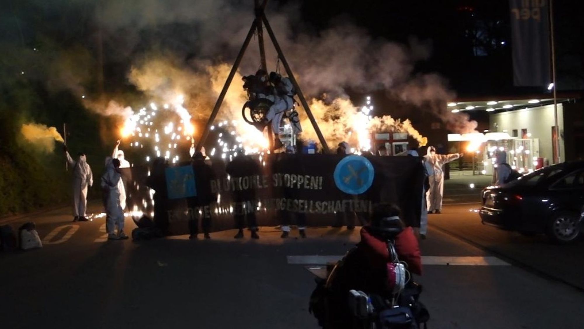 Menschen hängen in einem Dreibein , eine der 2 Personen im Rolltuhl. es ist Nacht. die Demonstrierenden tragen weiße Anzüge und haben Rauchtöpfe angezündet, der Rauch davon ist im Bild. auf dem Banner vor dem Tripod steht Blutkohle stoppen Energiekonzerne vergesellschaften und das Logo von Ende Gelände. im Vordergrund eine Peron im E-Rollstuhl. Das Bild wurde mit Langzeitbelichtung aufgenommen.