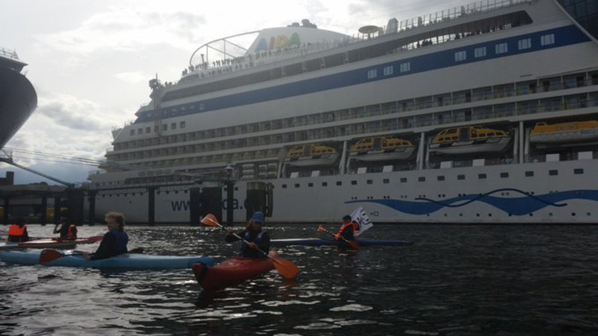 5 Kayaks im Wasser. Im Hintergrund ist ein kreuzfahrtschiff zu sehen.