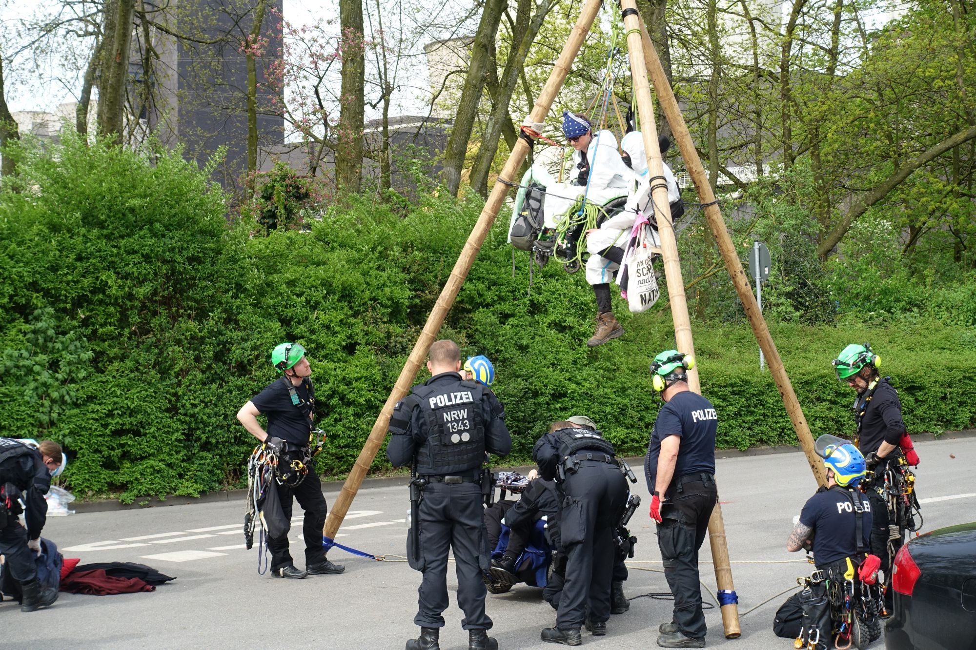 Polizeieinheiten mit und ohne Kletterausrüstung stehen am Tripod. Im Tripod 2 Menschen in Maleranzügen. Unten ein demonstranten im e-Rollstuhl, polizisten versuchen auf ihn einzureden.