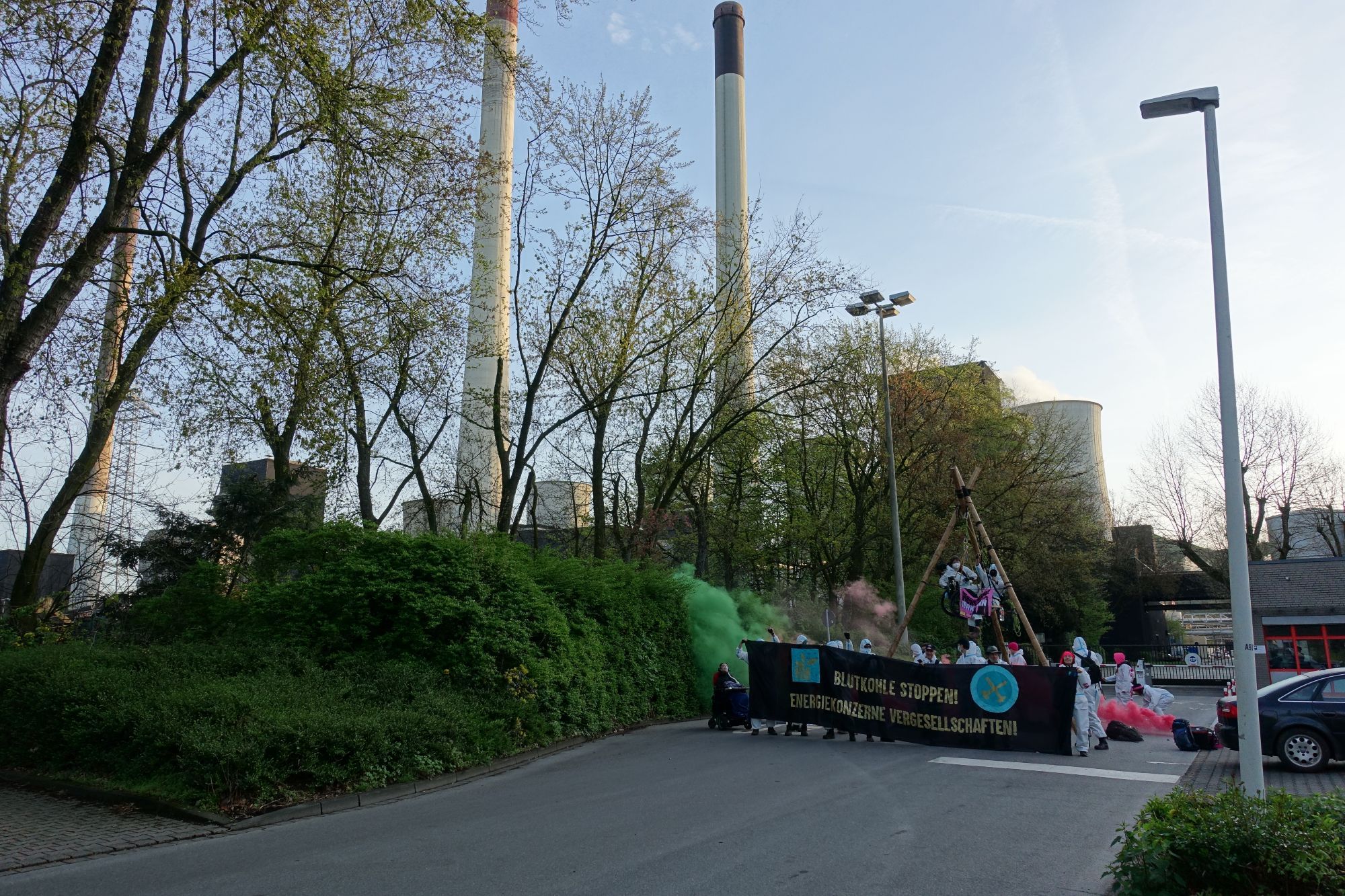 es ist inzwischen hell geworden. Aktion mit Tripod, 2 Menschen darin, Menschen mit weißen Maleranzügen und banner Blutkohle stoppen energiekonzerne vergesellschaften und grüner Rauch und das Kraftwerk im Hintergrund