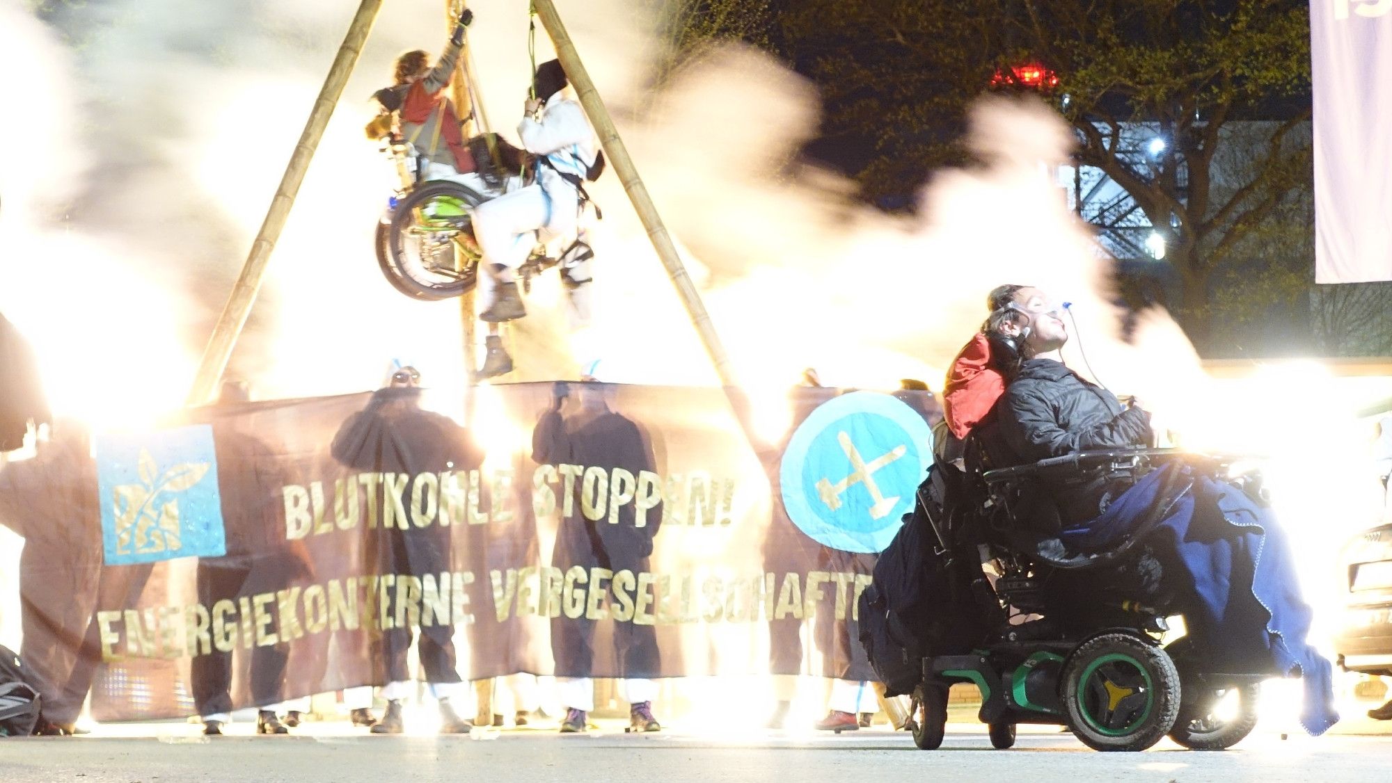 Menschen hängen in einem Dreibein , eine der 2 Personen im Rolltuhl. es ist Nacht. die Demonstrierenden tragen weiße Anzüge und haben Rauchtöpfe angezündet, der Rauch davon ist im Bild. auf dem Banner vor dem Tripod steht Blutkohle stoppen Energiekonzerne vergesellschaften und das Logo von Ende Gelände. im Vordergrund eine Peron im E-Rollstuhl. Das Bild wurde mit Langzeitbelichtung aufgenommen.