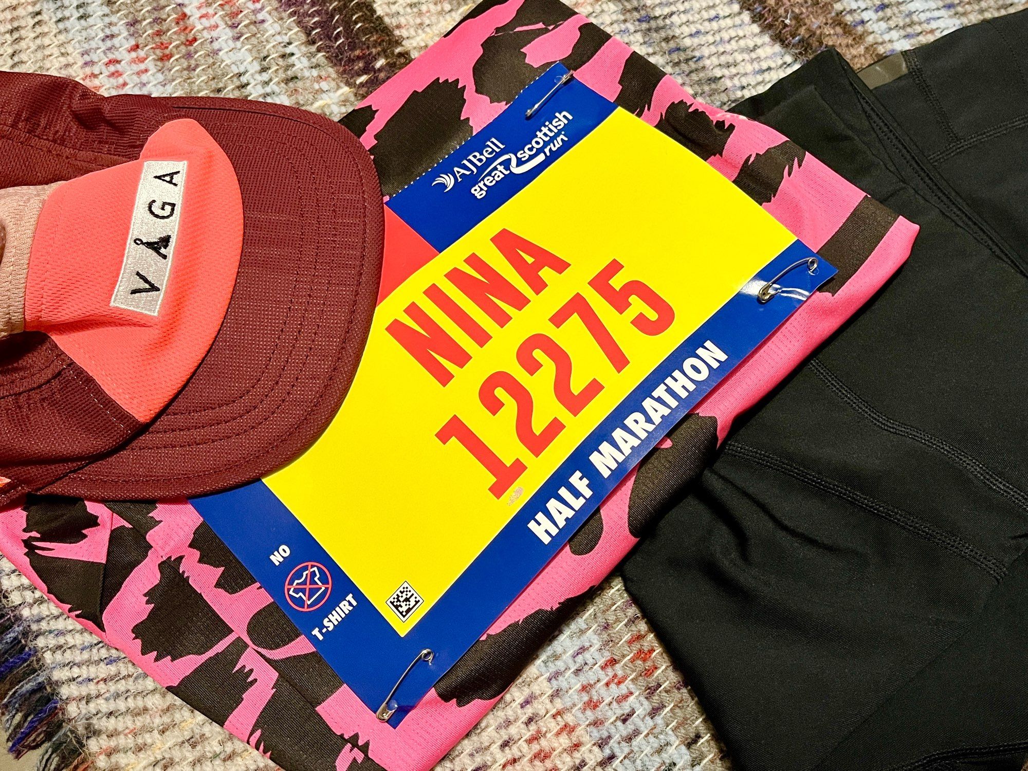 A flat lay of some running kit (a pink running cap, pink and black leopard print top, and black shorts) on a bed. The race number attached to the top is yellow, and in red letters says “NINA 12275”.