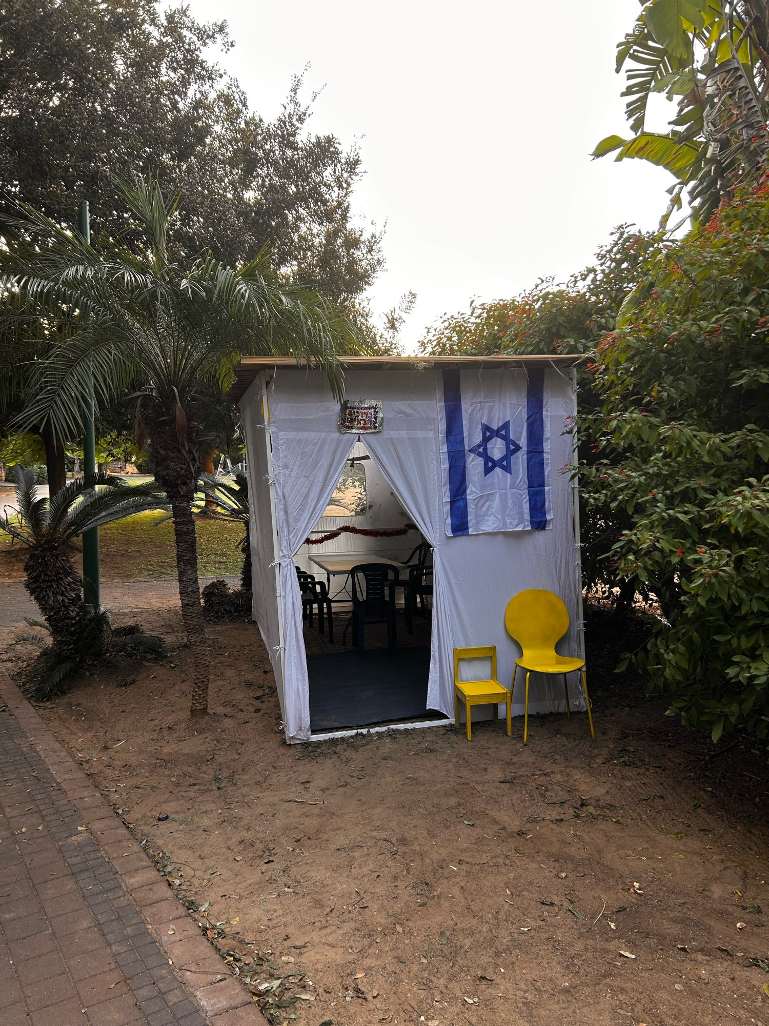 Eine Laubhütte von außen mit Wänden aus weißem Stoff. Neben dem Eingang stehen zwei gelbe Stühle, ein kleiner und ein großer. Darüber hängt die Fahne Israels. 