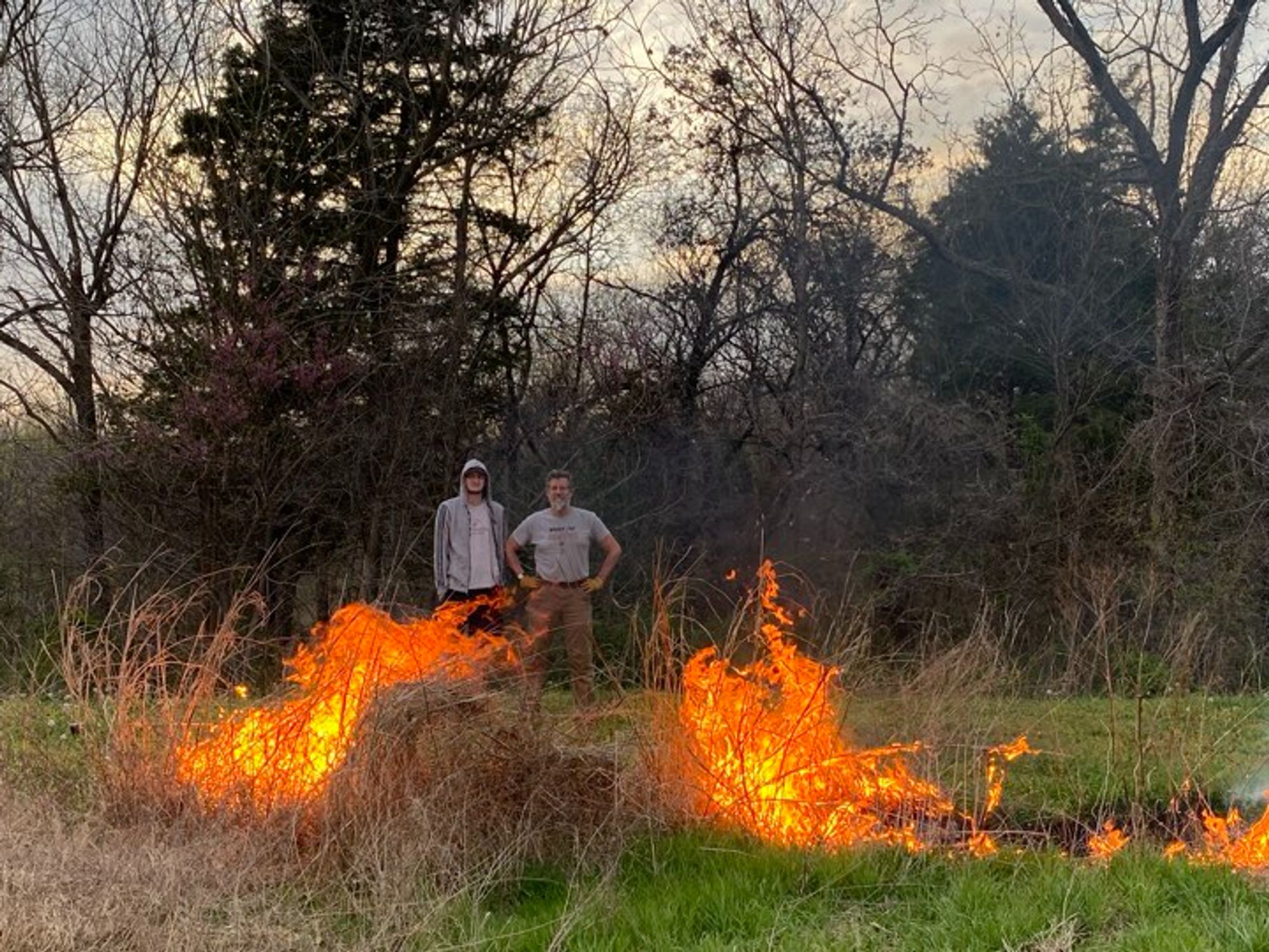 Two white guys standing behind a burning patch of last year's vegetation on a bright green lawn. The younger and taller one is wearing a gray hoodie. His dad is sucking in his gut but is unsuccessful in looking anymore youthful and trim for the effort.