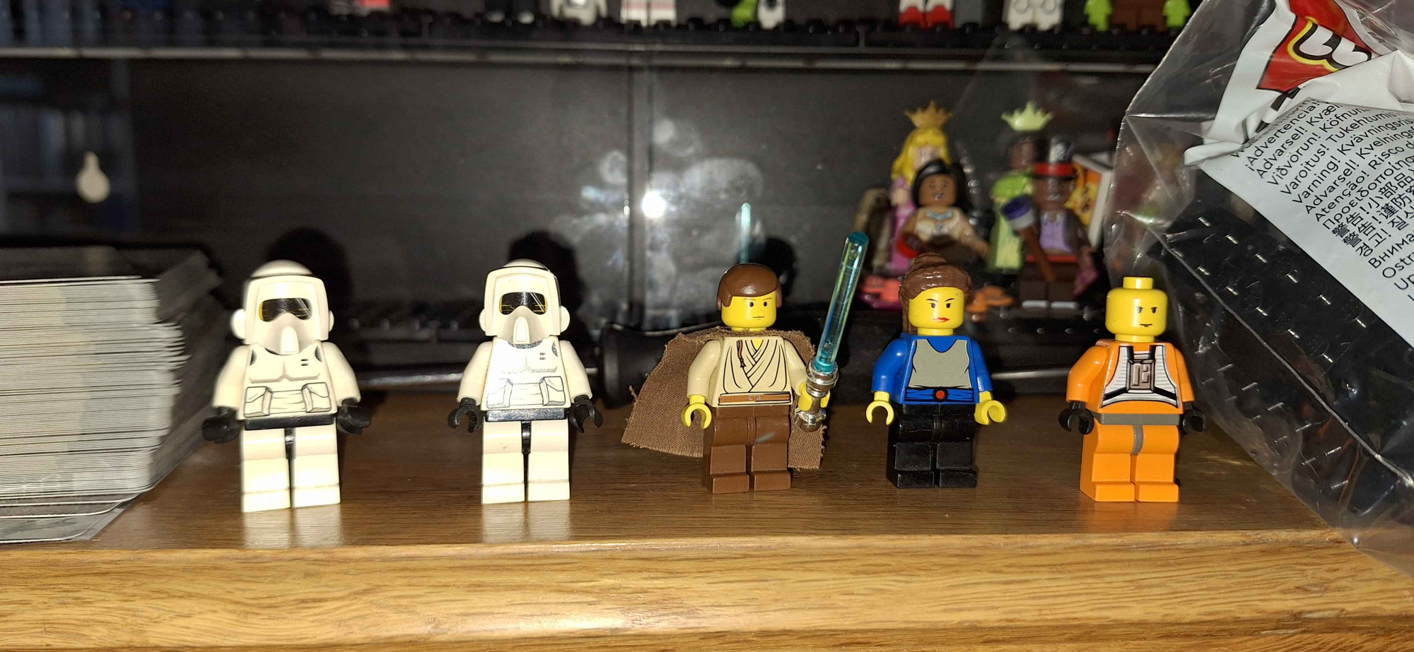 Five lego minifigures are standing side by side on top of a desk in front of display case. They are the original Lego Minifigures of two Stormtrooper Scouts, Phantom Menace Obi-Wan Kenobi, Admidala and Luke Skywalker as X-Wing pilot (without hair or helmet)