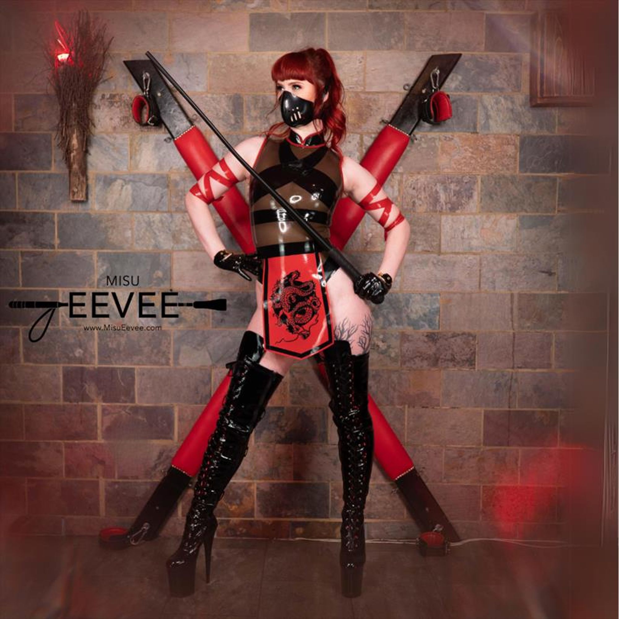 The image features a woman standing confidently in a bold, dominatrix-inspired outfit. She is wearing a black and red latex ensemble that includes a high-neck top, gloves, and thigh-high lace-up boots. A striking red and black front panel features a dragon design, adding a warrior aesthetic. She is also wearing a black latex face mask and holding a long whip in one hand, exuding a powerful, commanding presence. Behind her is a large red and black St. Andrew's cross attached to a stone wall, further enhancing the dominatrix theme. The setting has a moody, dungeon-like atmosphere with dim lighting, and there are red accents on the walls, including a red light.. The overall look blends elements of fetish fashion and samurai warrior aesthetics.