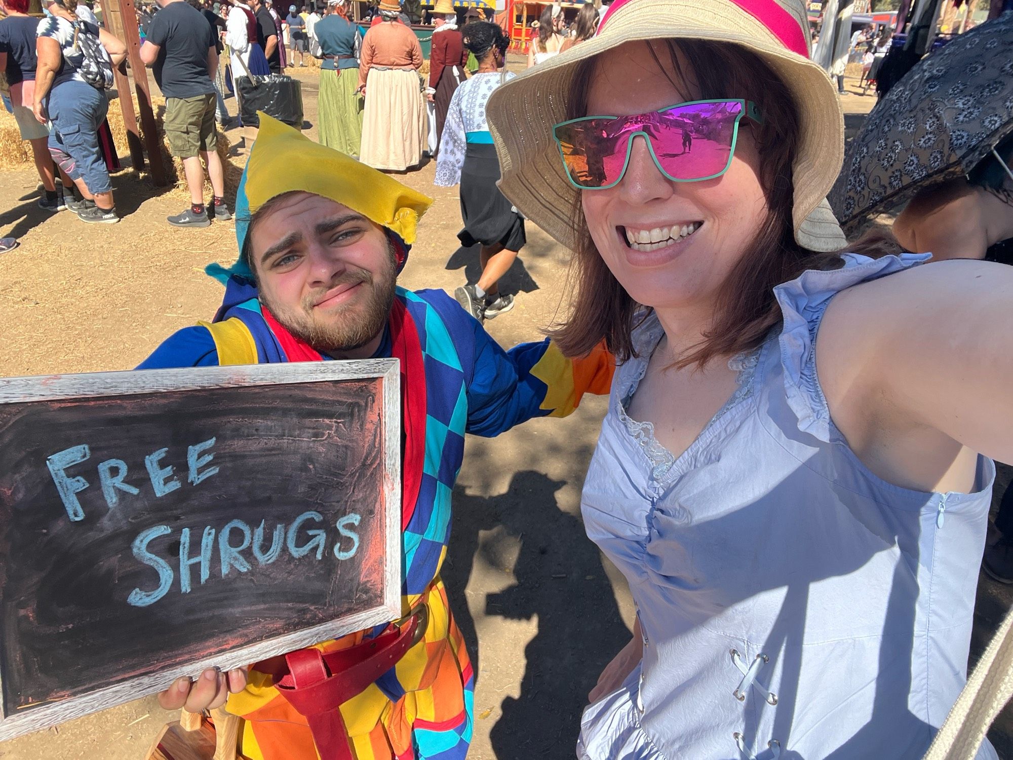 Holly, in an olde timey dress, next to a jester shrugging while holding a "free shrugs" sign at a Renaissance faire