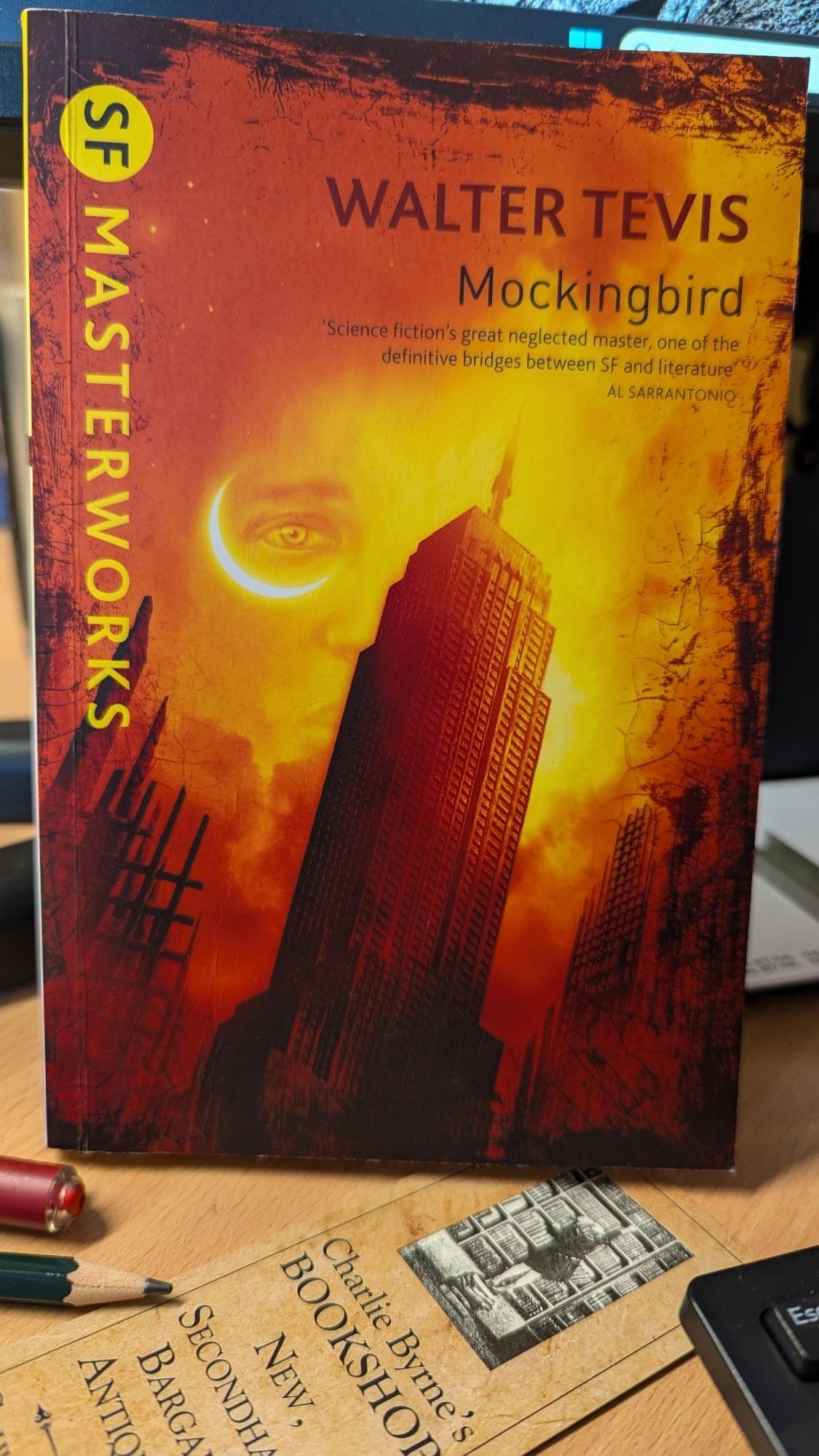 The cover of the SF Masterworks paperback edition of the book 'Mockingbird' by Walter Tevis. It shows an imposing view of the Empire State Building from below against a background in various shades of orange and the suggestion of a face in the background, with the right eye encircled by a crescent moon.