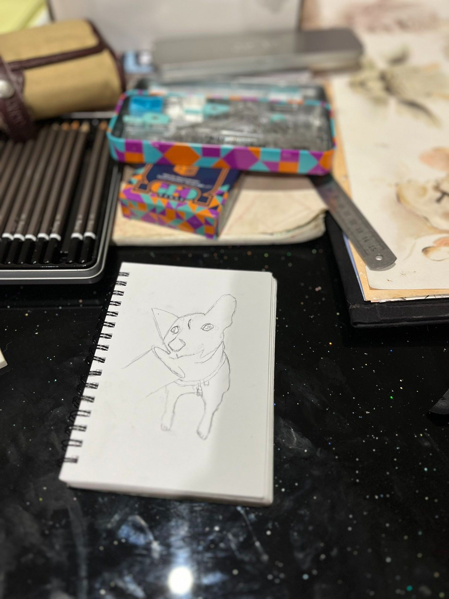 A very rough sketch of a dog sitting, looking at the viewer. He belongs to the relatively unknown subspecies Canis lupus familiaris subsp. puppuccinius. The sketch is drawn in pencil on a spiral-bound notebook. Various art supplies, including pencils, rulers, and sketchbooks, are visible in the background.
