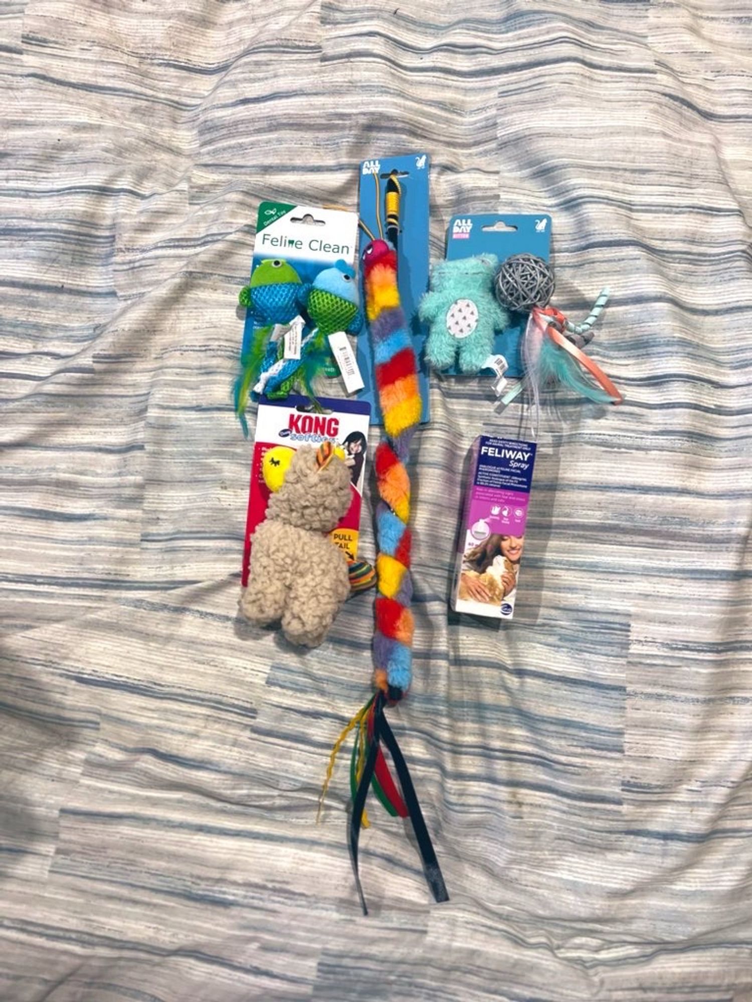 A collection of cat toys sitting on a bed. There is a rainbow caterpillar on a stick, a cute little light blue frog toy, a wicker ball with feathers, a vibrating llama toy, two dental fish toys and a spray bottle of Feliway.