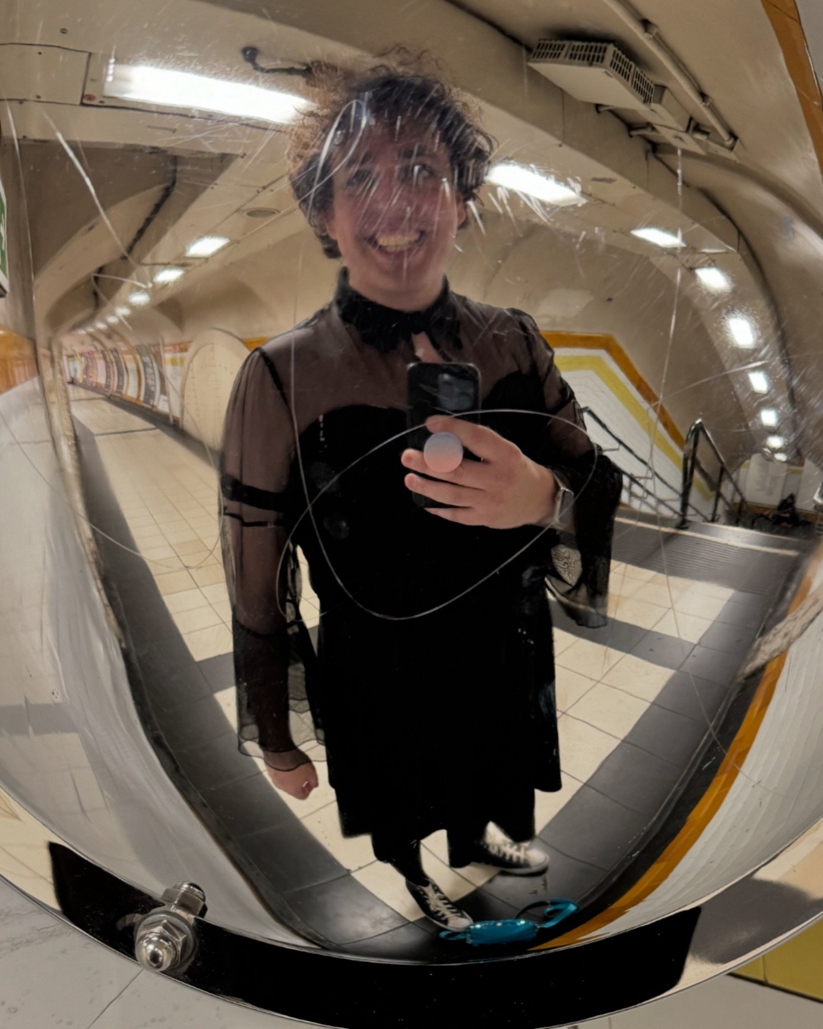 Selfie taken in a convex mirror in a tube station corridor. I have an absolutely beaming grin, wearing a a black dress with collar, and sheer sleeves and shoulders. It’s a little witchy I suppose.