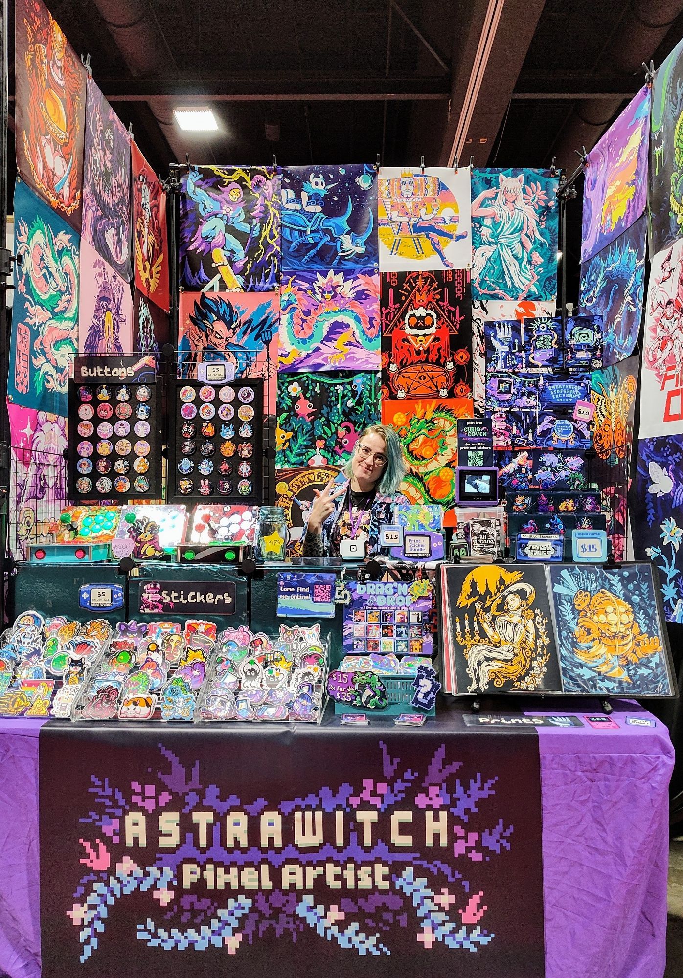 My pixel art booth setup, me standing behind the table with large bright prints behind