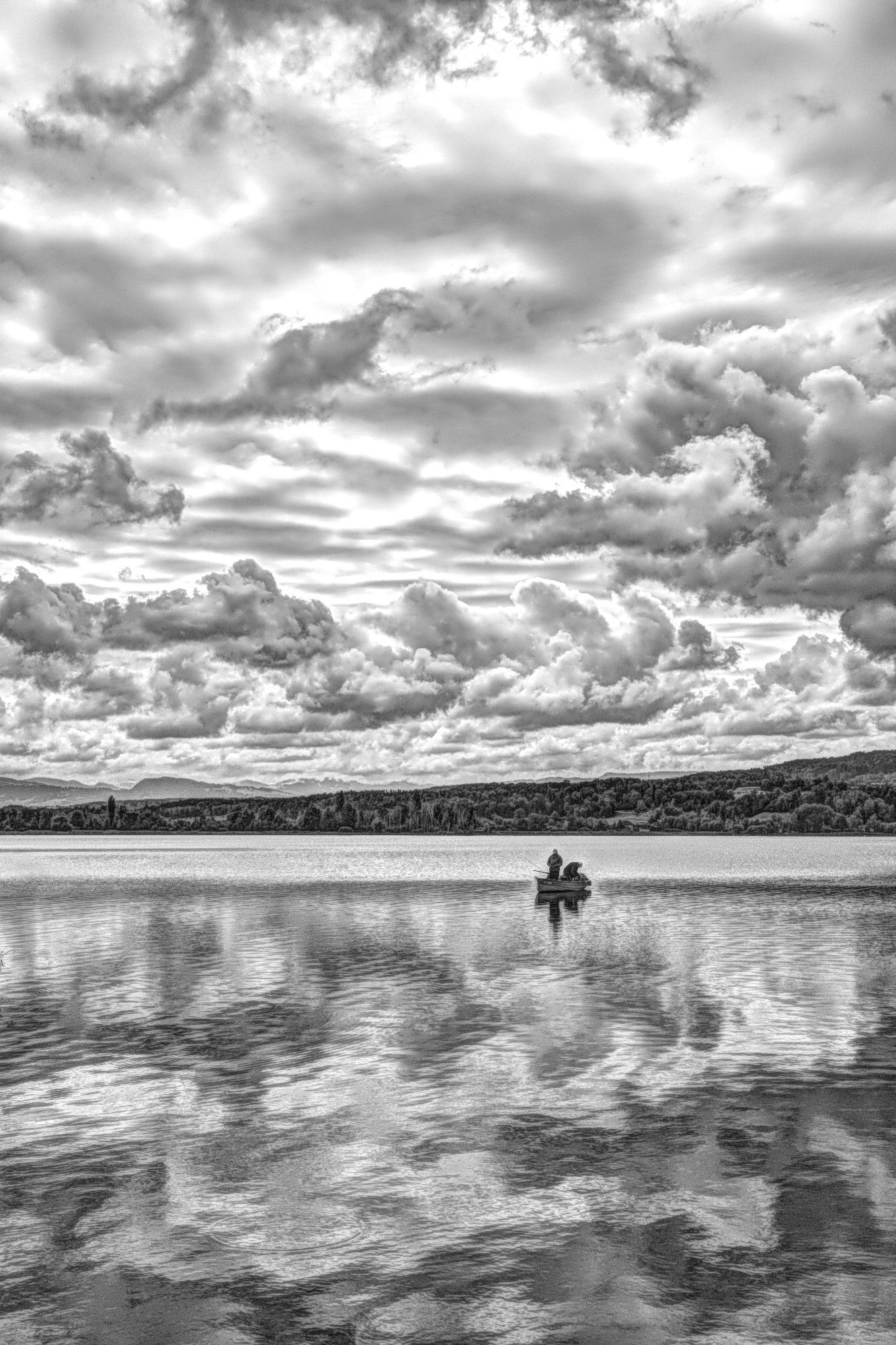 Fotografie in HDR Schwarzweiß mit einer Ricoh GRIIIx. Man erkennt im Vordergrund einen See und am Himmel eine schöne Wolkenszene. Am zweiten Blick erkennt ein Fischerboot mit zwei Personen.