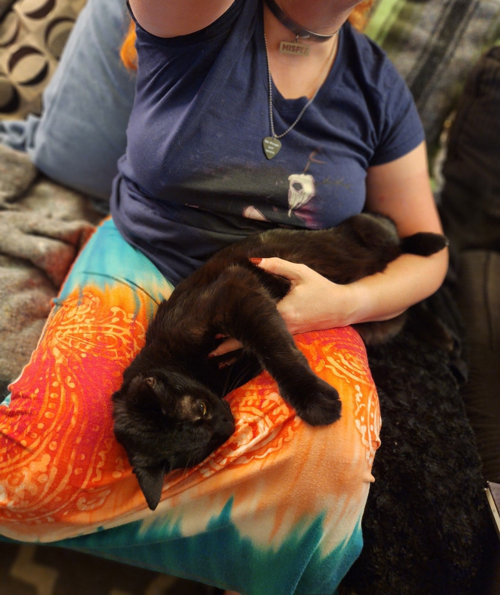 Woman wearing a long skirt, holding a black cat with one arm on her lap.