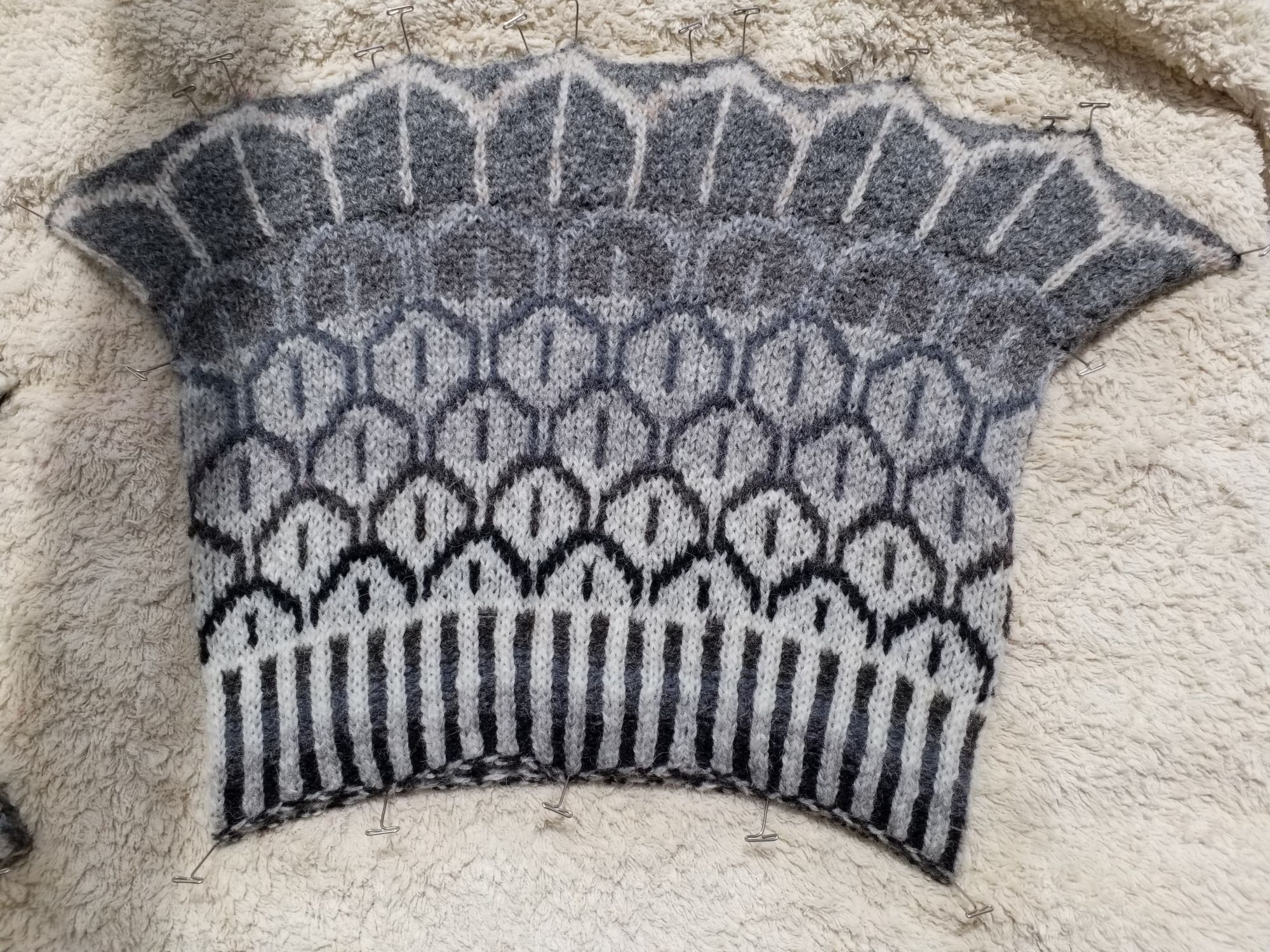 A cowl blocking, featuring a deep corrugated rib and a stylised feather pattern. Worked in shades of grey, the cowl is flared to sit on the shoulders