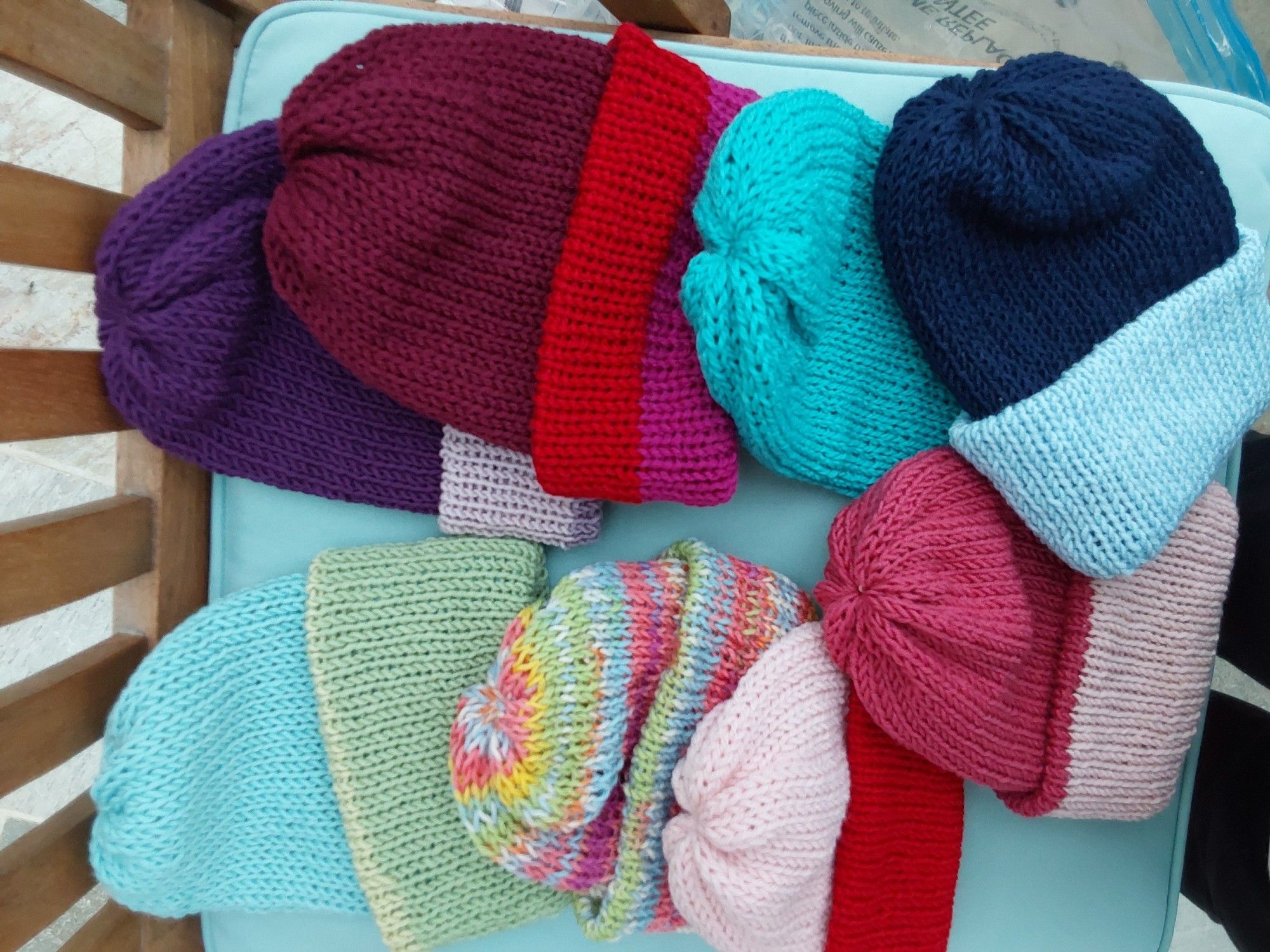 The hats are double thick with a gathered crown and folded brim. They are lying on a chair. Top row: purple and light purple, 3 shades of red, turquoise, dark blue and light blue.
Bottom row: Light blue and light green, multicoloured striped self-patterning, red and pink, 2 shades of pink.